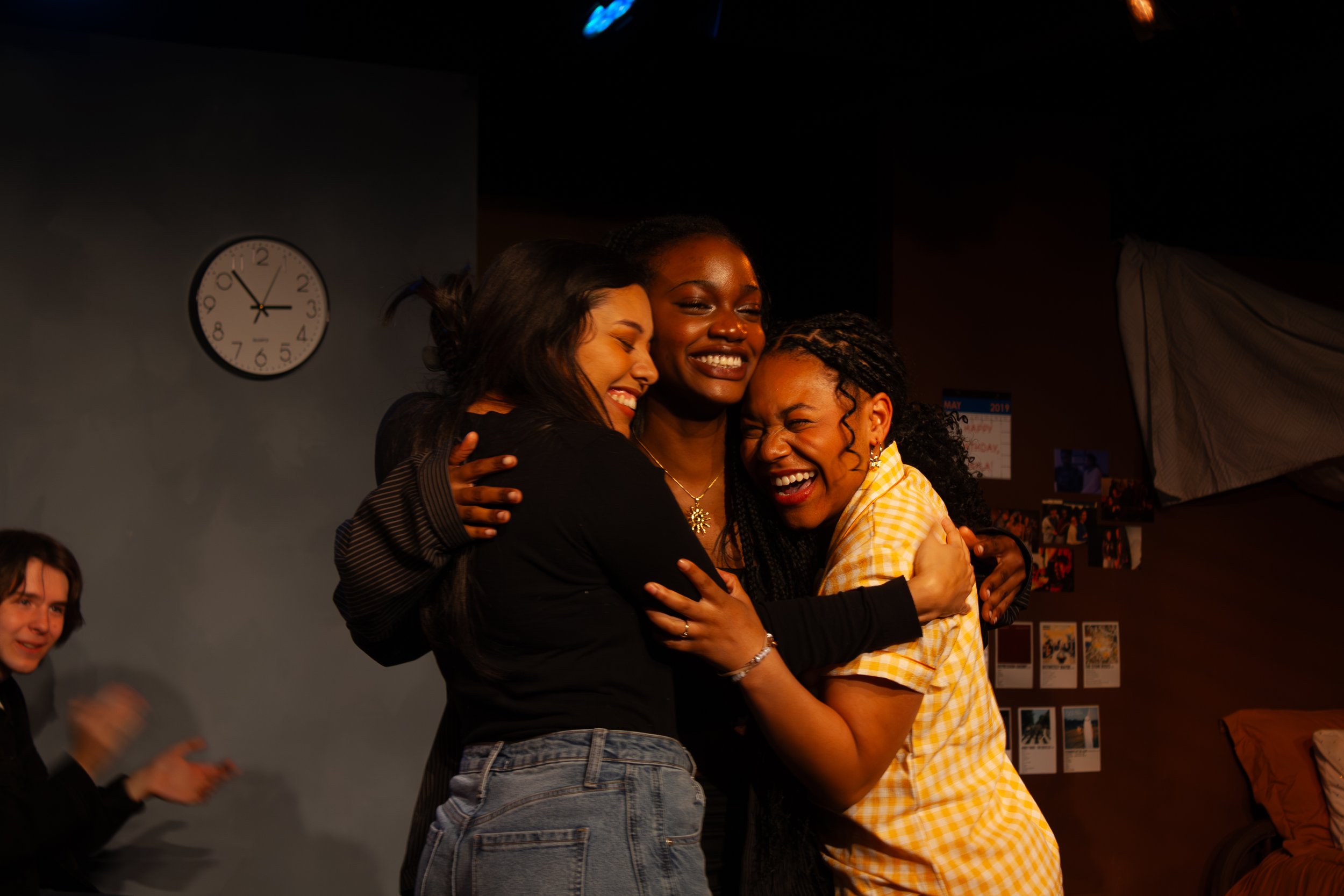 Three women hugging and smiling at a gathering or celebration, with a clock on the wall showing 2:46 and a calendar on the wall in the background.