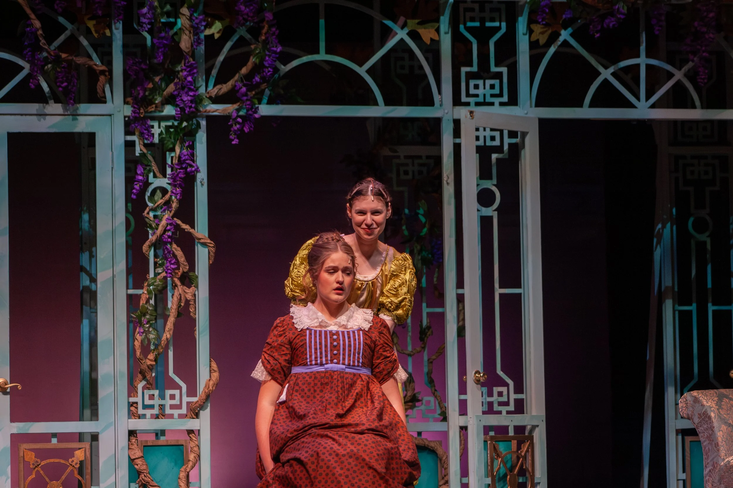 Stage scene depicting three women in period costumes, with architectural and floral set design, in a theatrical performance.