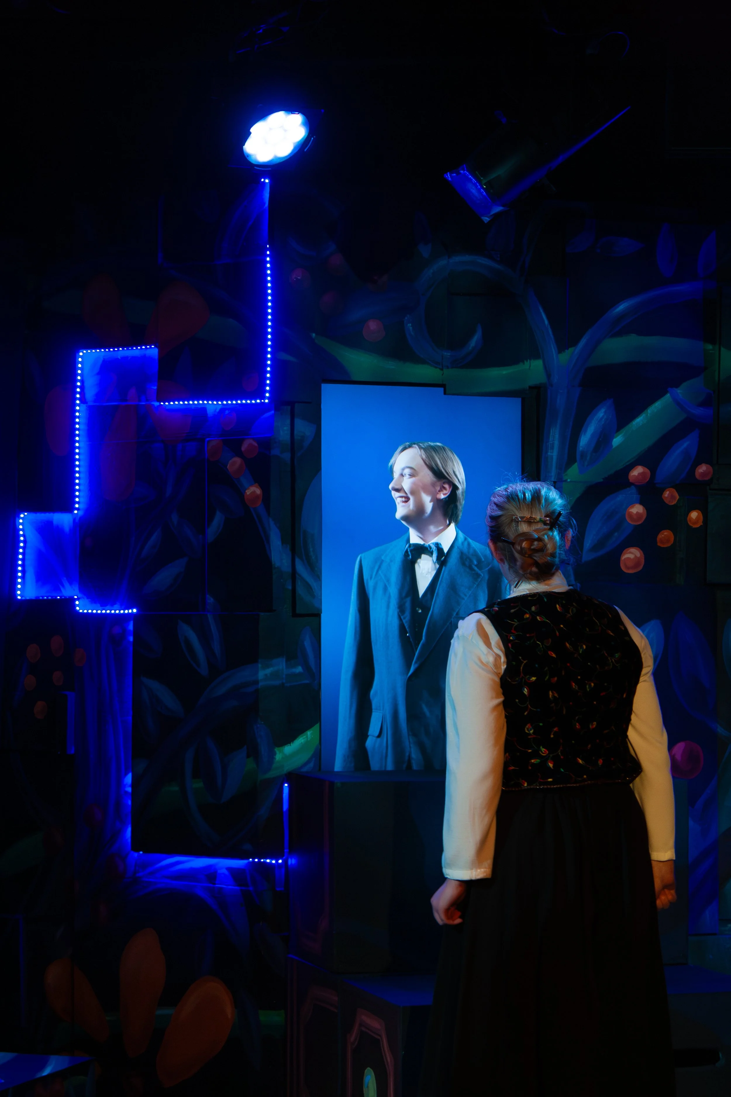 A brightly lit portrait of a woman in a suit and tie, displayed on a large screen, with a person in the foreground viewing it in a dark, colorful room.
