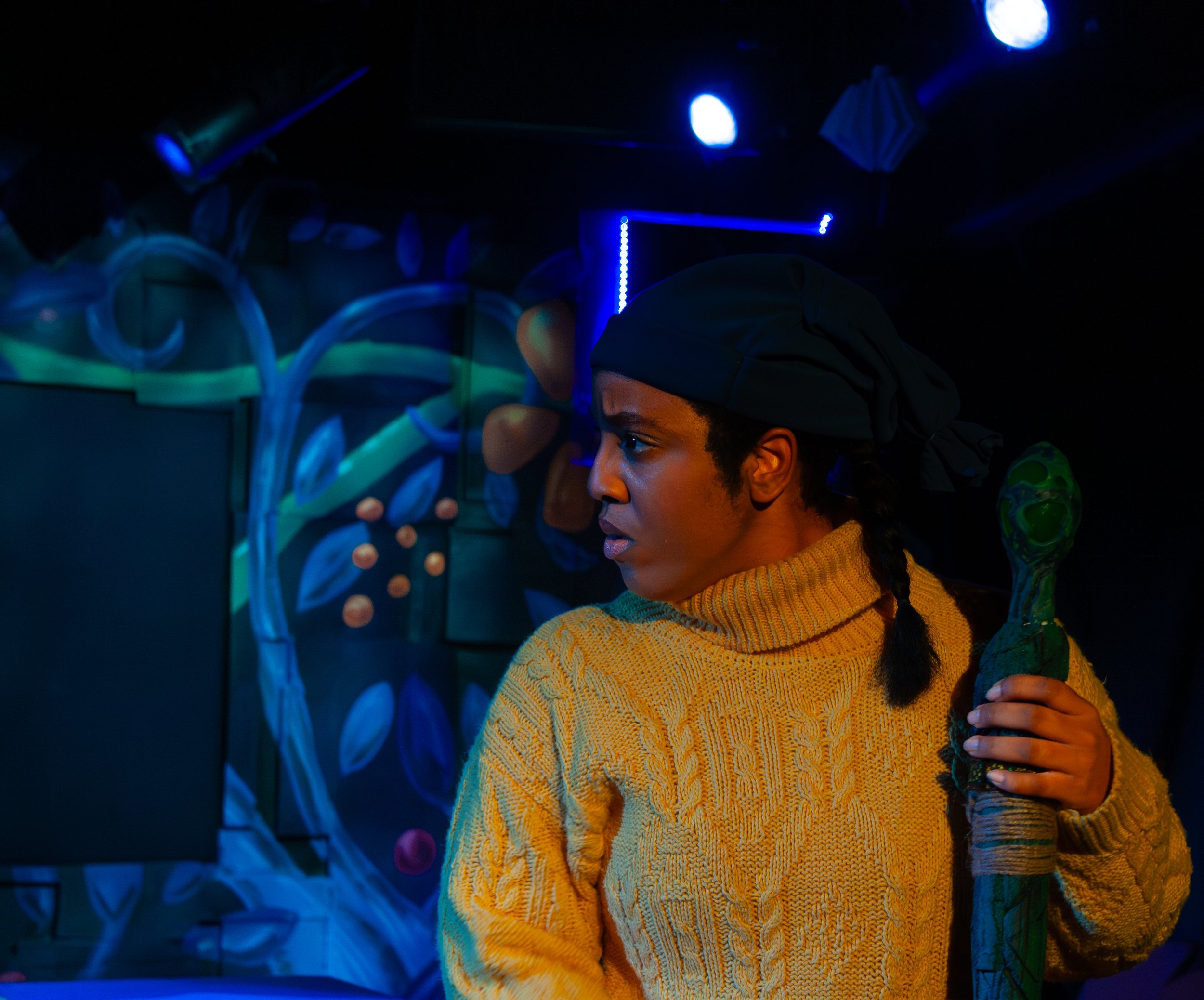 A person in a yellow sweater with braided hair is in a dark room with blue lighting, standing near colorful mural art and abstract decor.