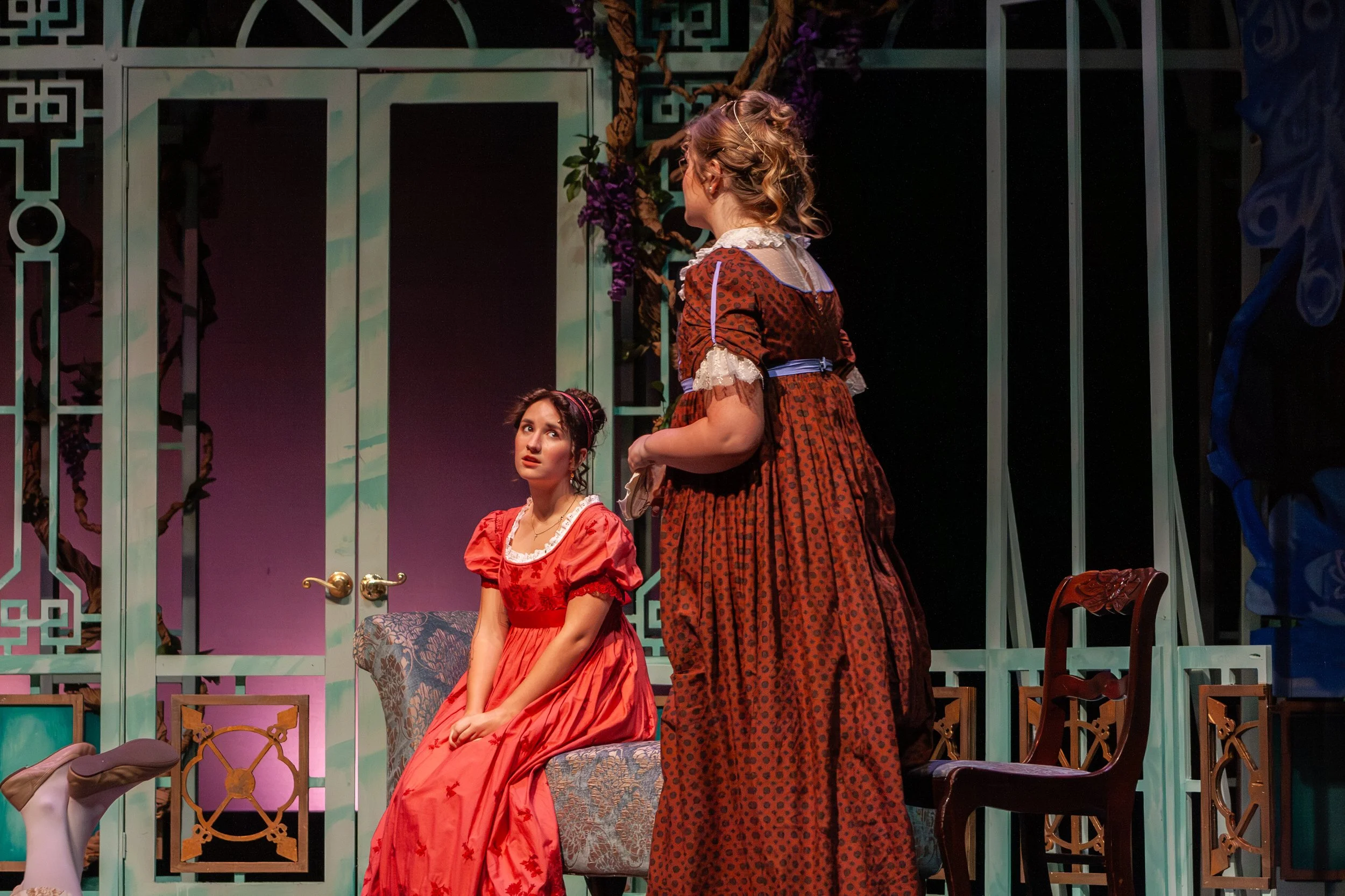 Two women in period costumes participating in a theatrical play. One woman is seated on a chair, wearing a red dress, looking up with a concerned expression. The other woman, standing, is wearing a patterned brown dress with lace trim, and she appear