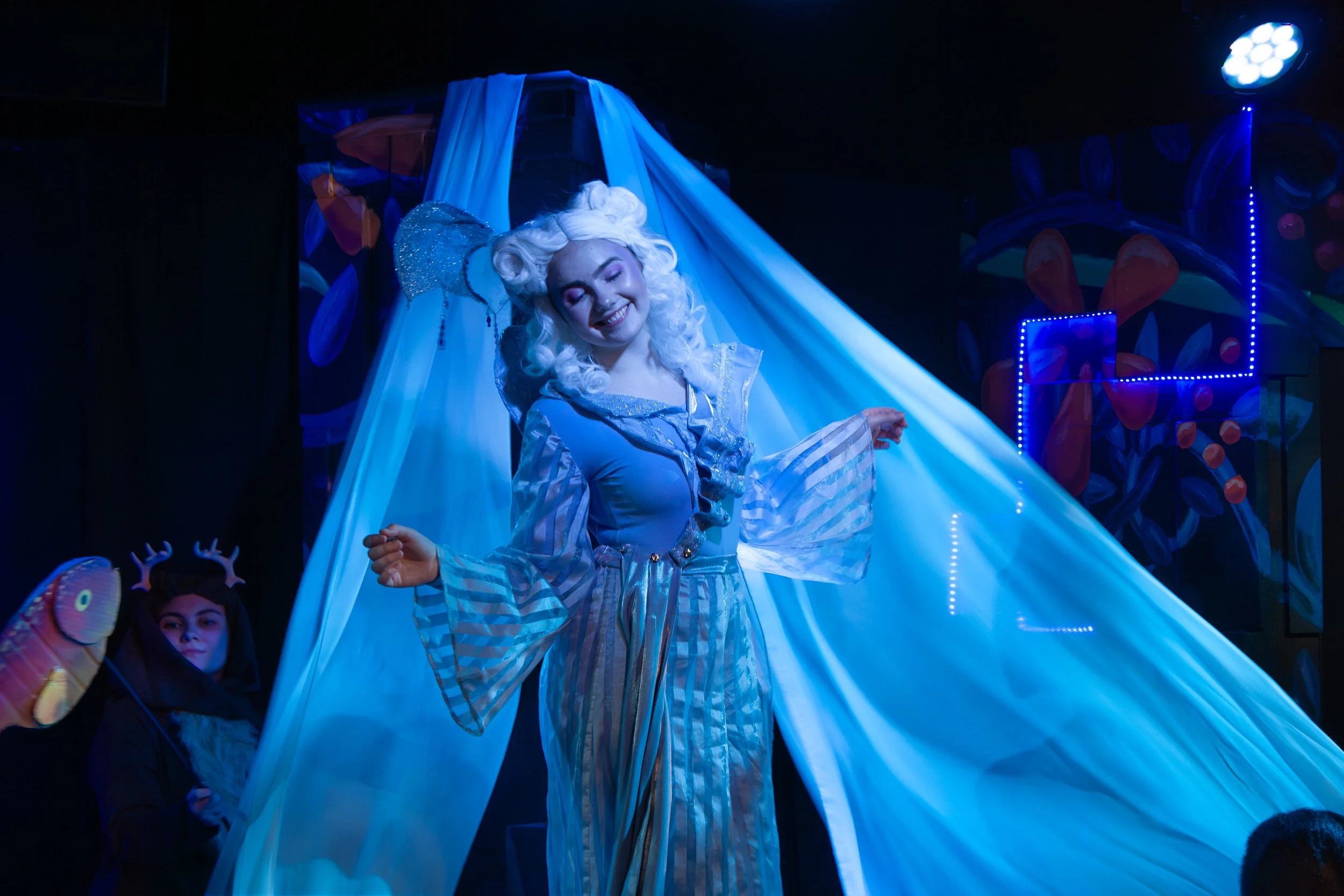 A performer dressed as a character from a fairy tale, with a white curly wig and a sparkly costume, dancing or entertaining in front of an audience in a dark setting with colorful stage lights.
