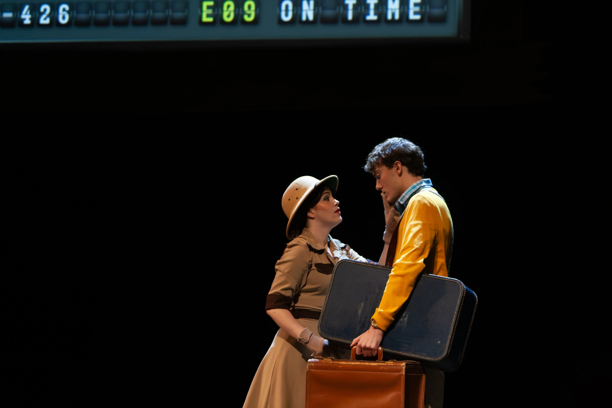A male actor in a yellow jacket and a female actor in a brown dress with a hat facing each other on stage, surrounded by chairs, with a dark background and text on a live scoreboard above them.