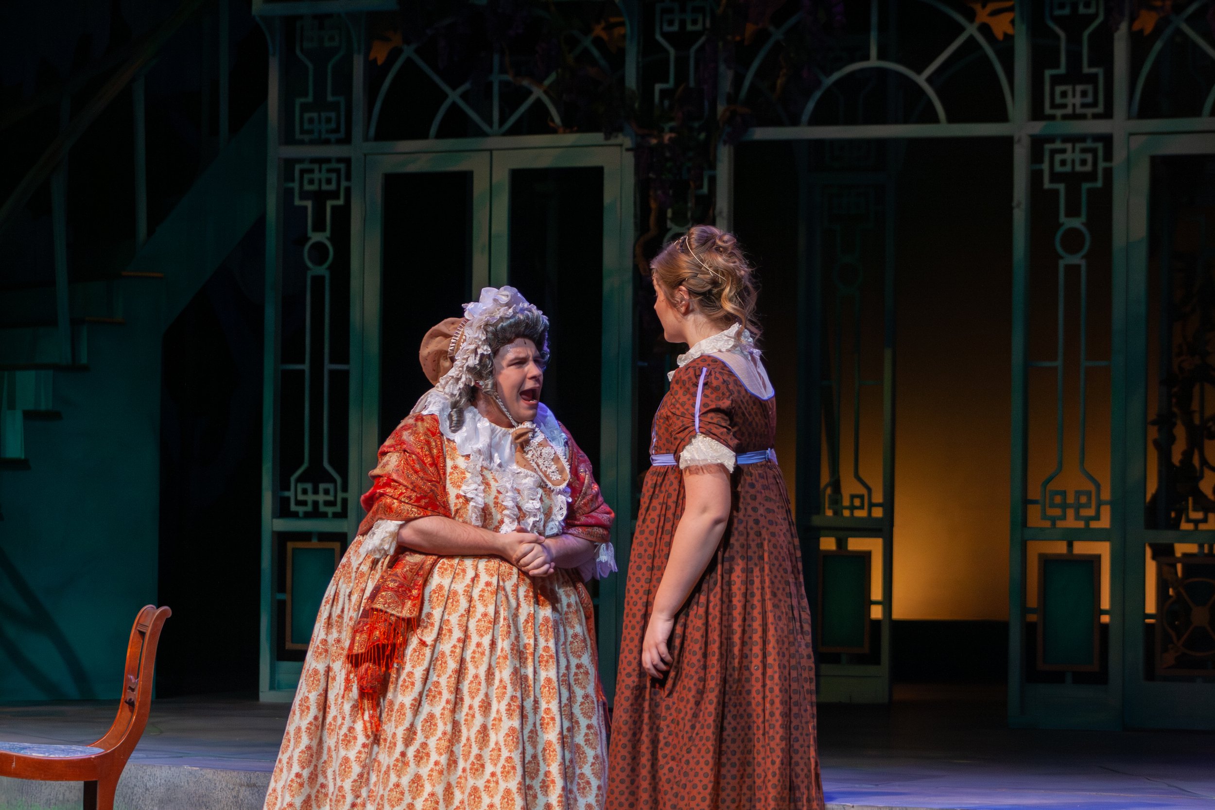 Two women in historical costumes, one older and one younger, engaged in a conversation on stage with a decorative background, likely in a theater production.