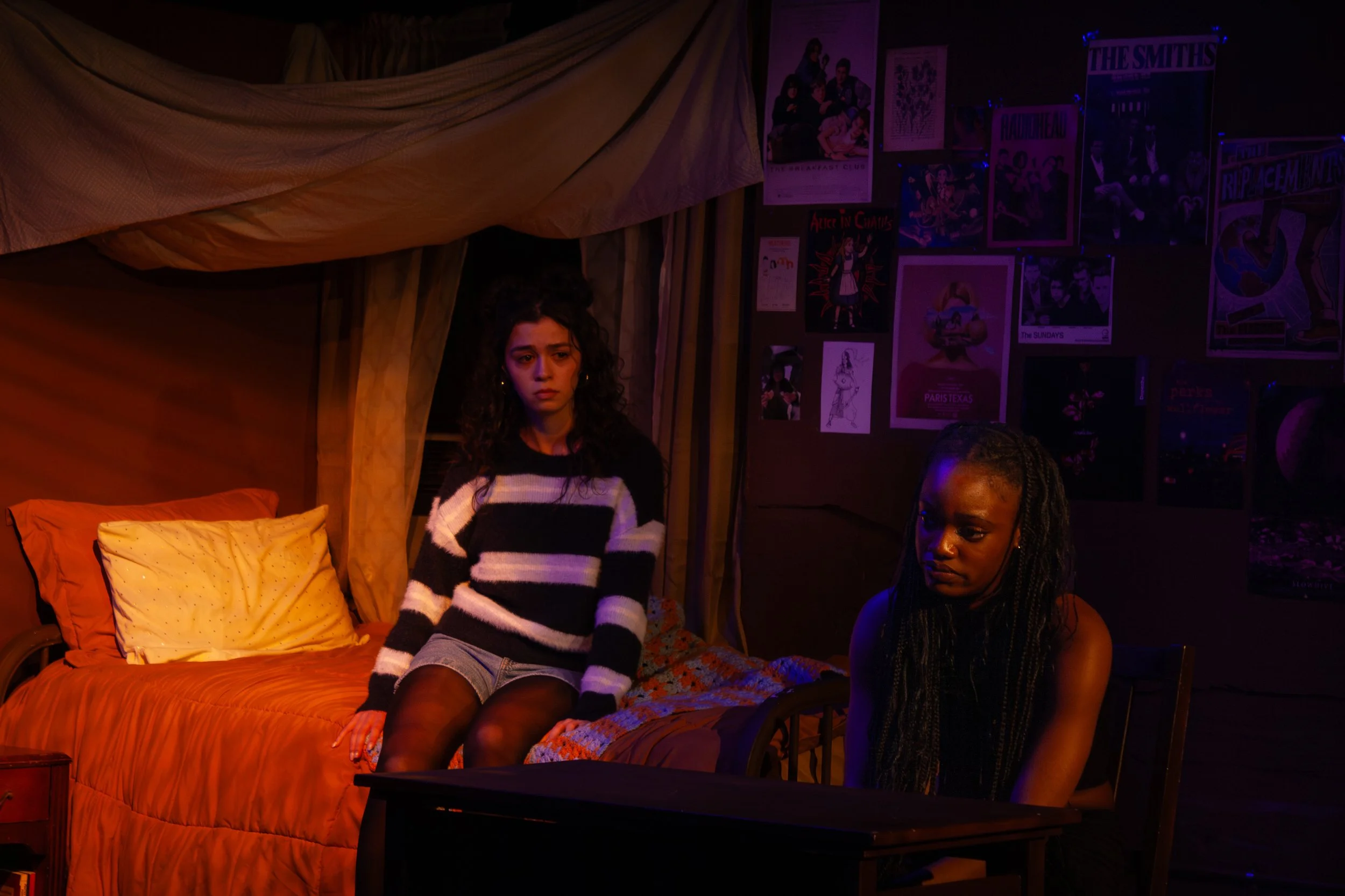 Two young women sitting on a bed in a dimly lit room decorated with posters and artwork on the walls. One woman has curly hair and is wearing a striped sweater, while the other has braided hair and is wearing a black tank top.