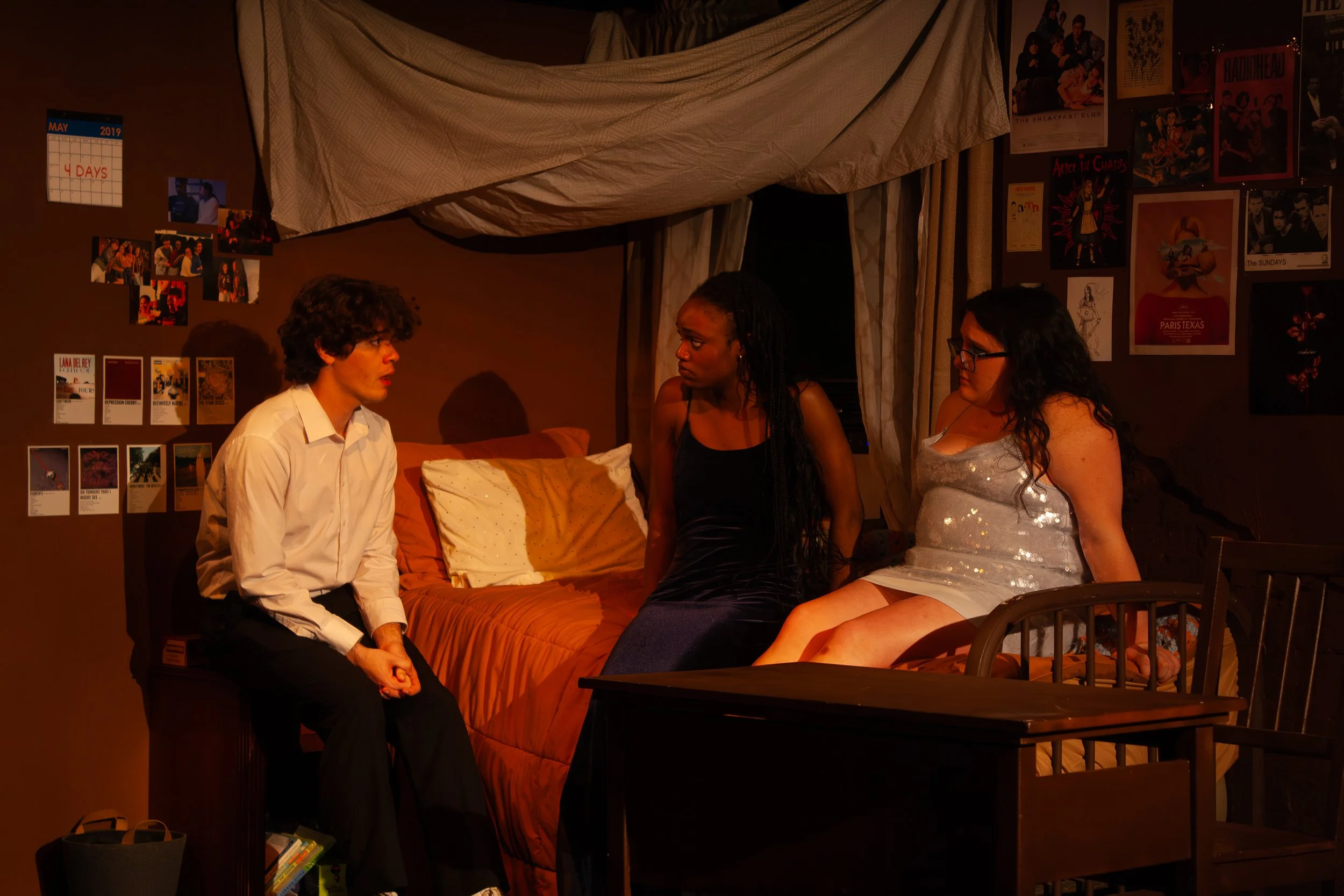 Three women and one man sitting in a dimly lit room decorated with movie posters and a calendar on the wall, engaged in conversation.