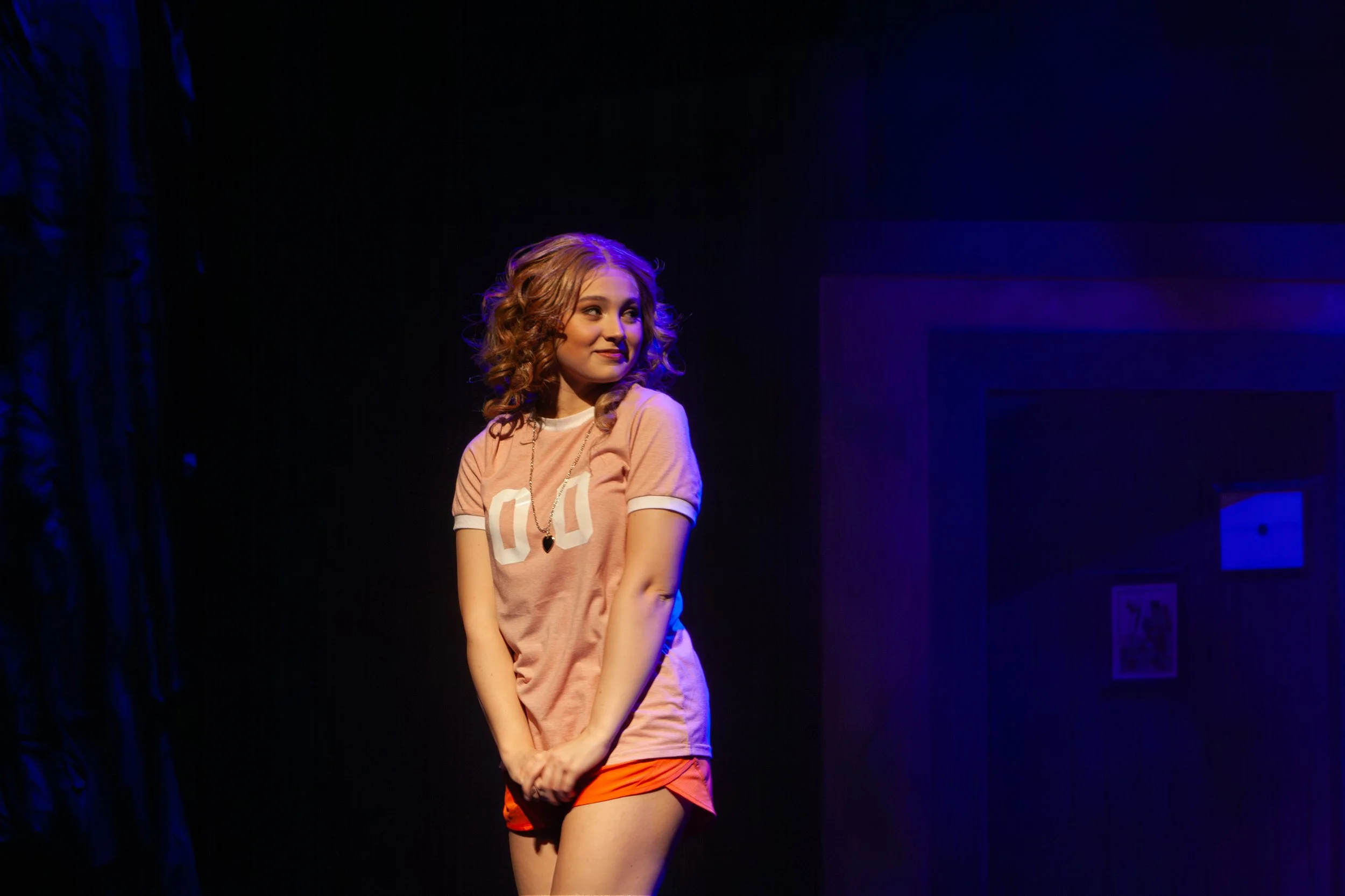 A young woman with curly hair wearing a pink t-shirt and orange shorts stands on stage under purple lighting, smiling and looking to her right.