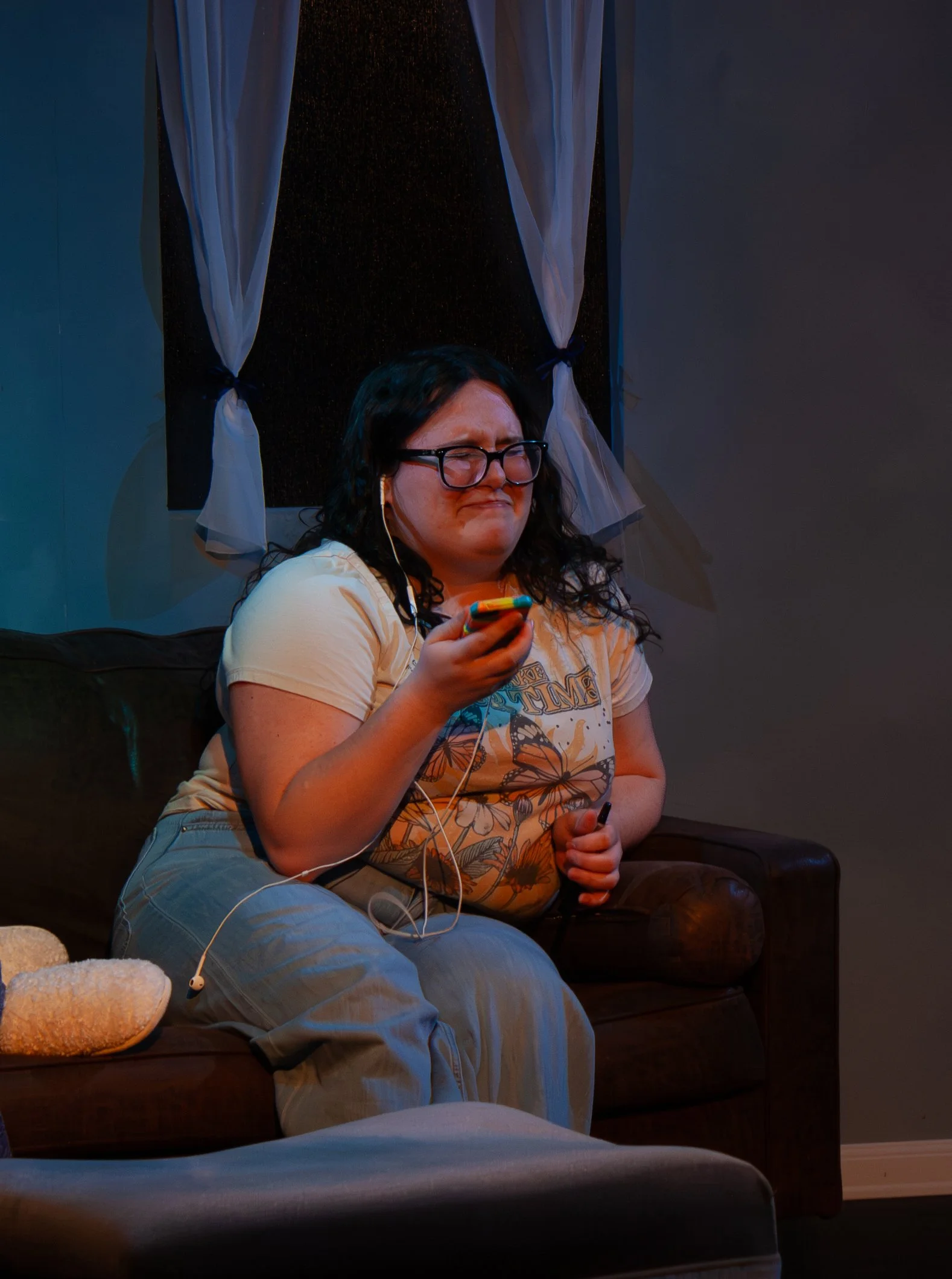 A woman with glasses and long dark hair sitting on a brown leather couch, smiling with her eyes closed while holding a smartphone. She is wearing a yellowish t-shirt with a graphic design, blue jeans, and white earbuds. There is a blanket or towel on