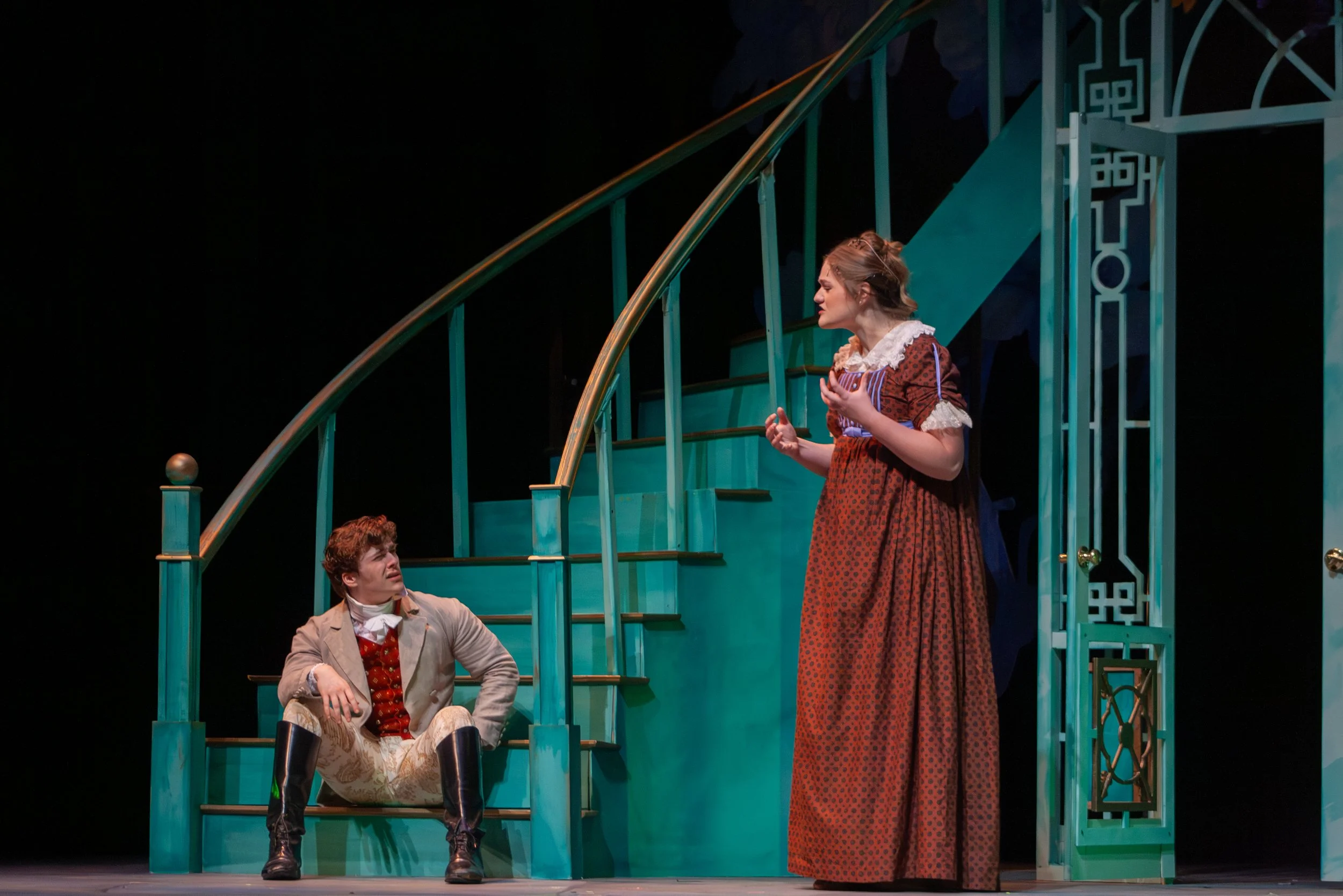 Three actors on stage performing in a theater production. Two are seated on stage, one man wearing a beige suit with a red vest and black boots, and a woman with an elaborate braid hairstyle wearing a long red patterned dress. The woman stands nearby
