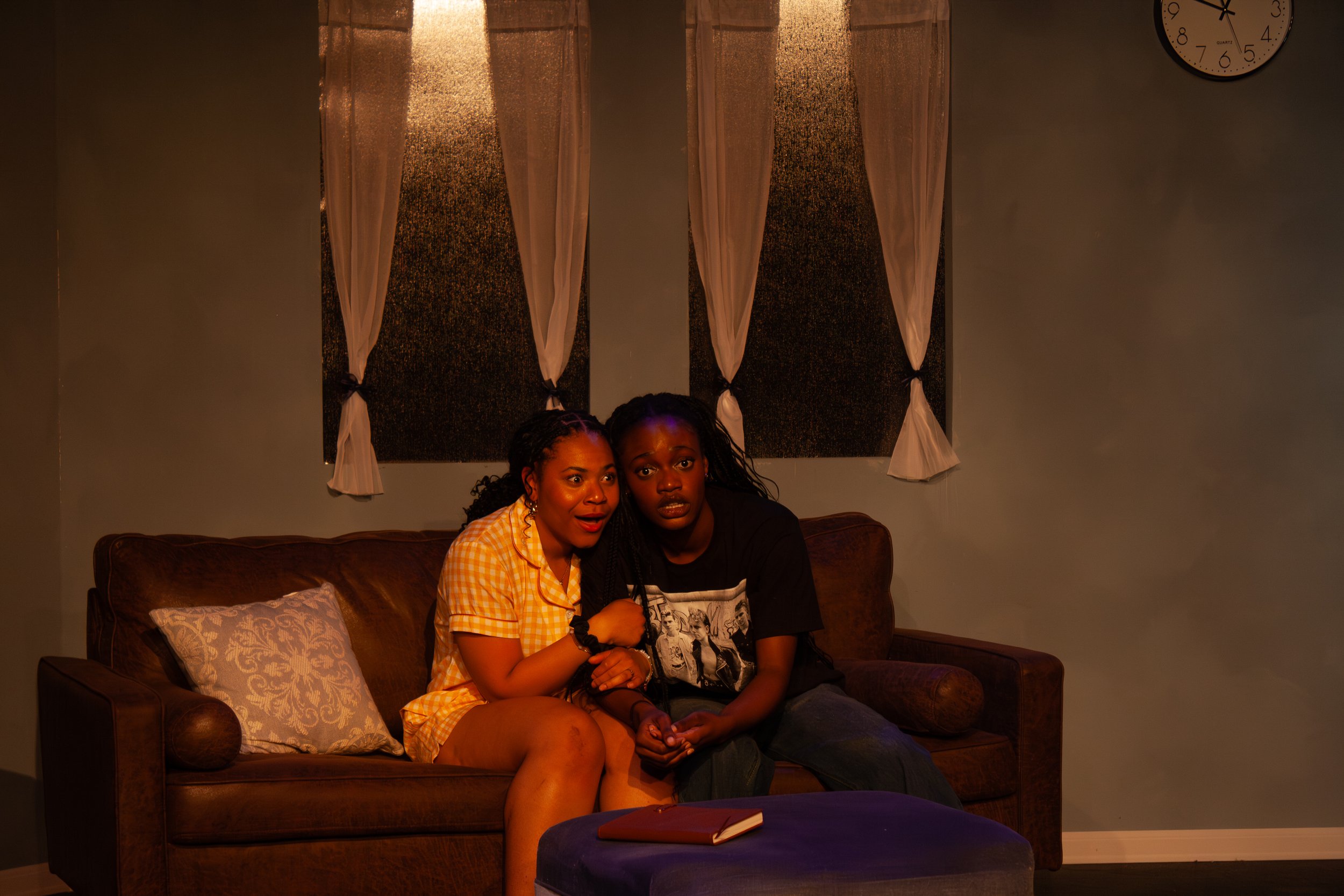 Two women sitting on a brown leather couch in a dimly lit room, one wearing a yellow checkered shirt and the other wearing a black T-shirt with graphic prints. They are close together, appearing surprised or shocked, with a red notebook on a purple o