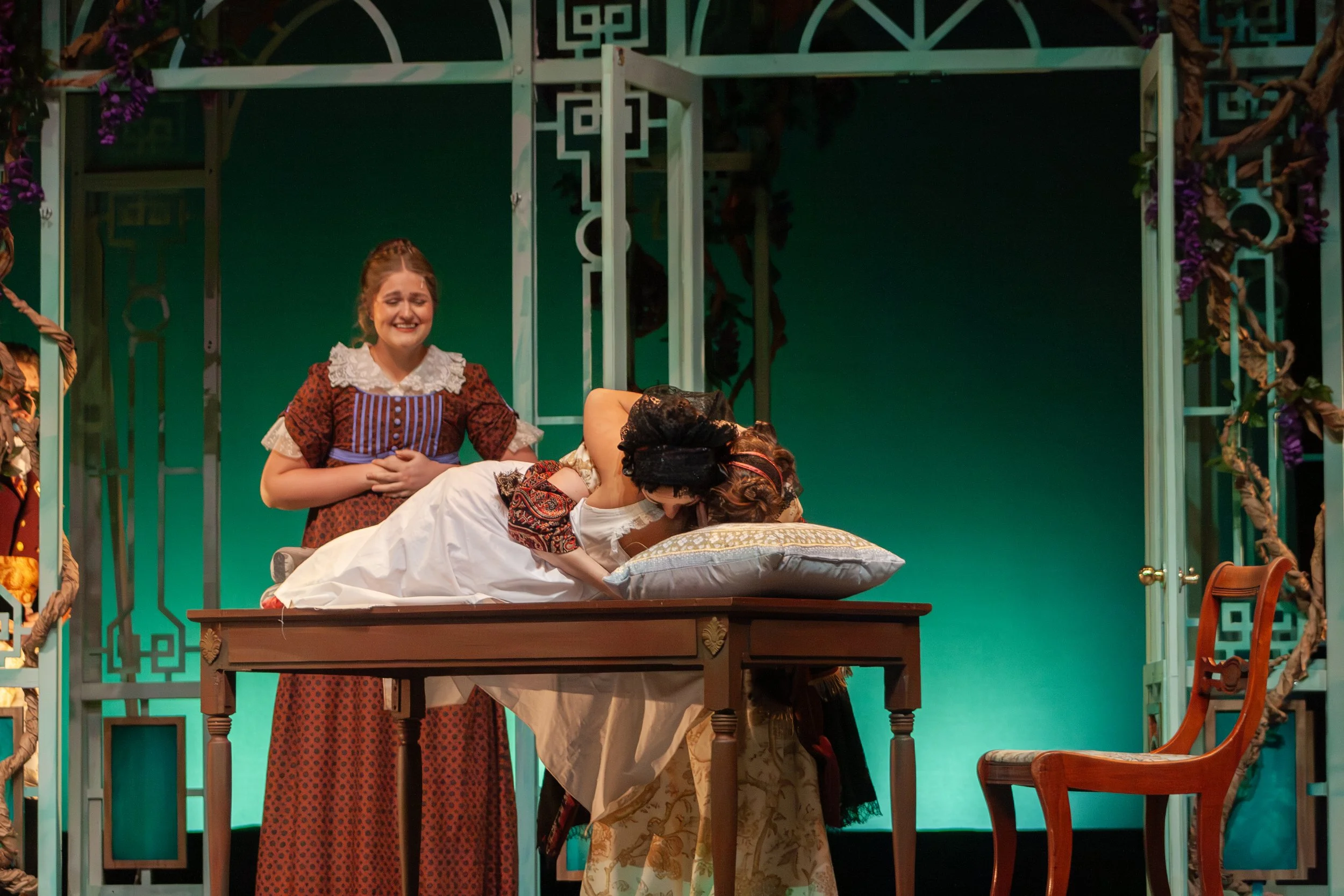 Two women in period costumes, one standing and smiling, the other lying on a bed, in a room decorated with vines and geometric patterns.
