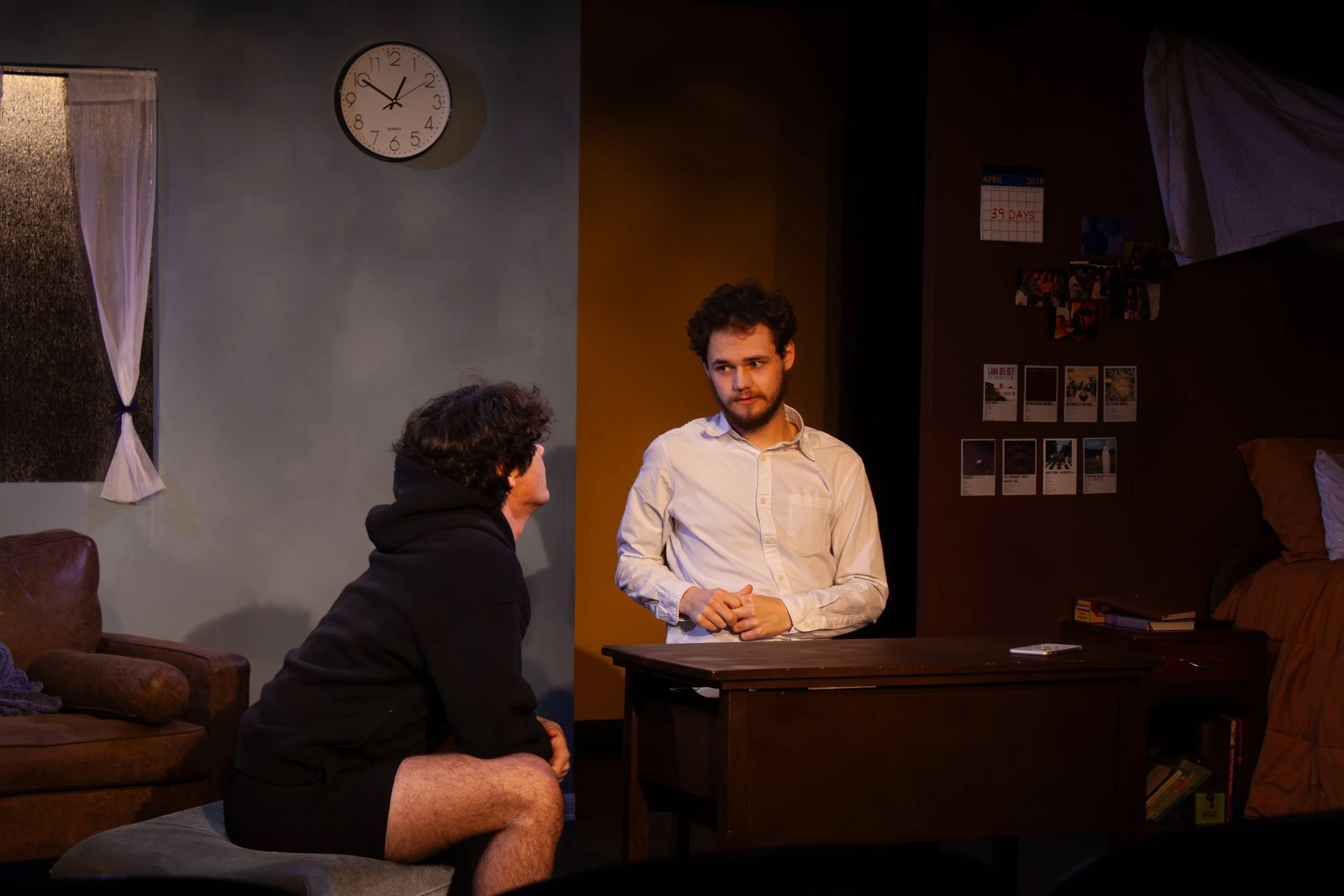 Two young men having a conversation in a dimly lit room. One is sitting on a couch with shorts and a hoodie, while the other stands by a table wearing a white shirt. On the wall behind them, there is a clock showing 9:07, a calendar indicating 39 day