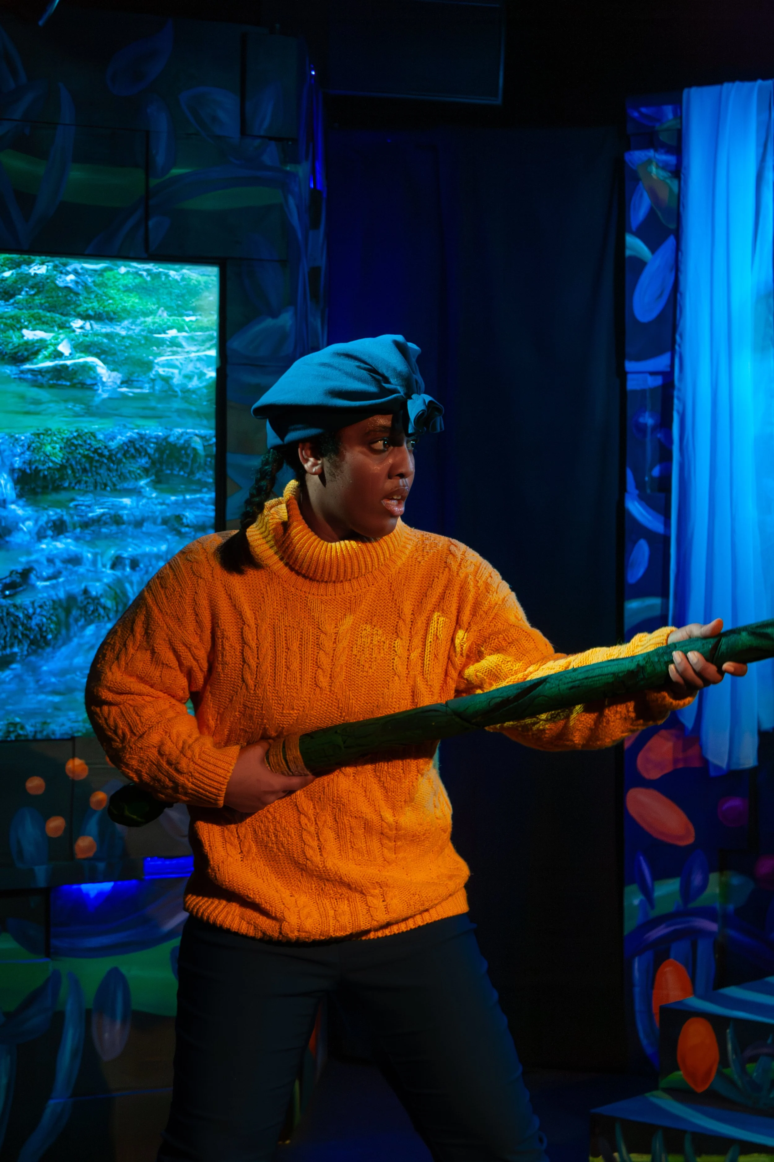A woman wearing a blue headscarf, orange sweater, and black pants, holding a large green stick, standing in front of a colorful mural with abstract patterns and a screen depicting a nature scene.