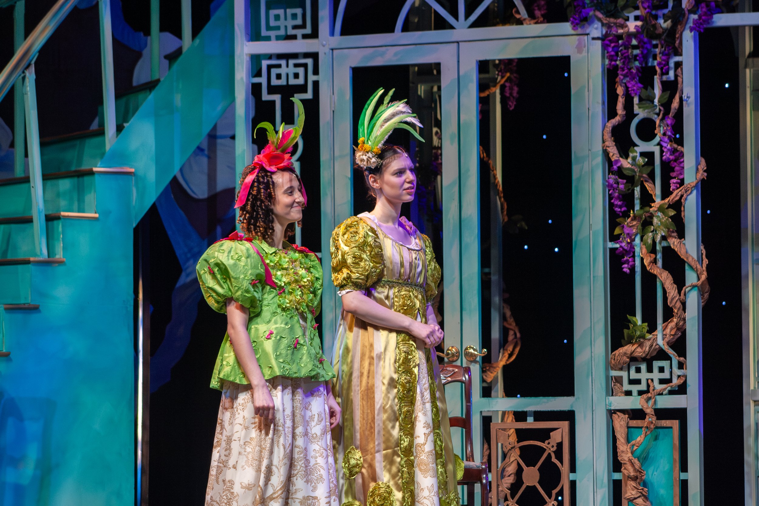 Two women in colorful costumes with floral and feather headpieces stand on a stage with decorative metal and plant elements.