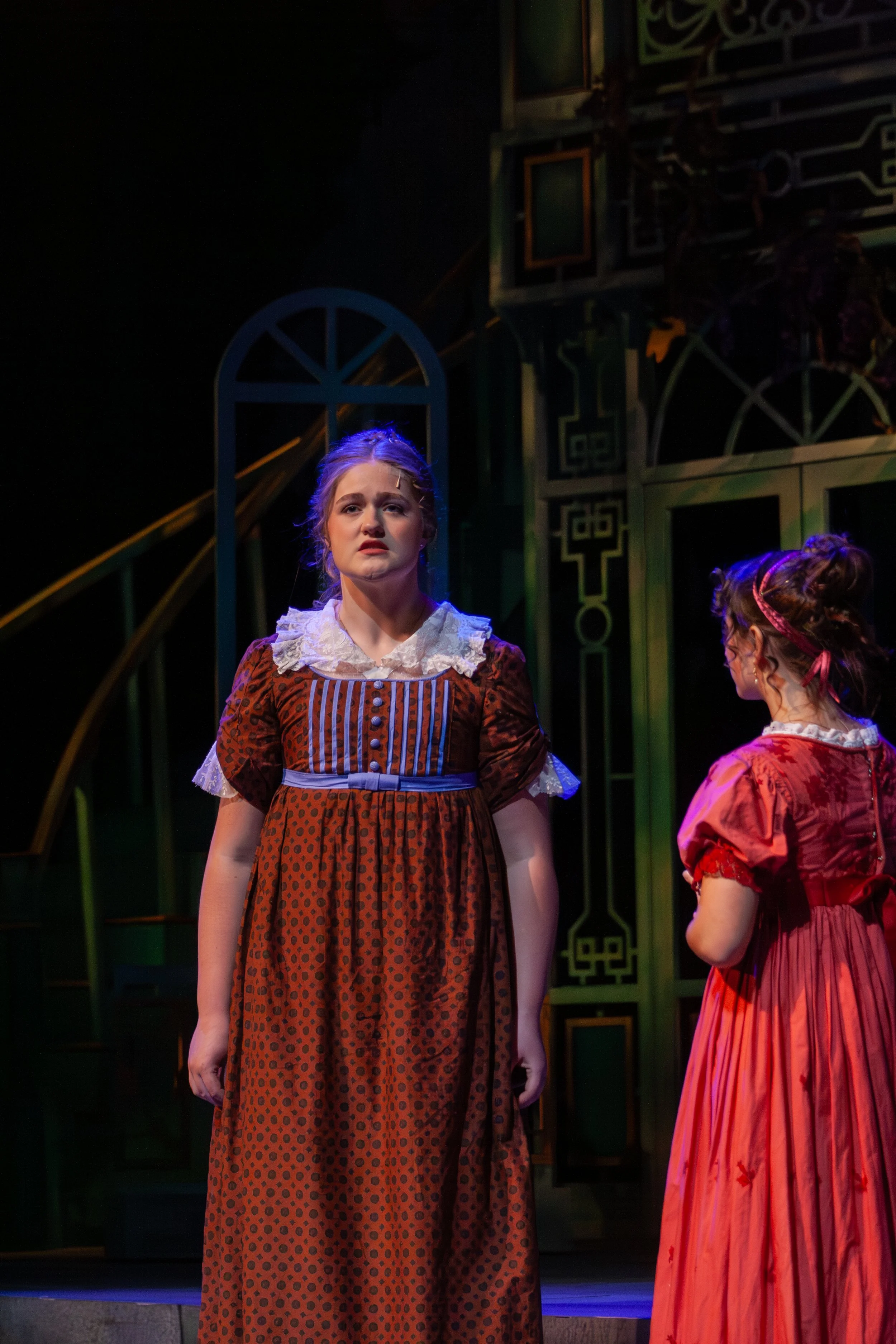 Two women in period costumes on stage, one in a brown dress with a white lace collar and the other in a pink dress with tree branch patterns, with a decorative green and gold set and arched window behind them.