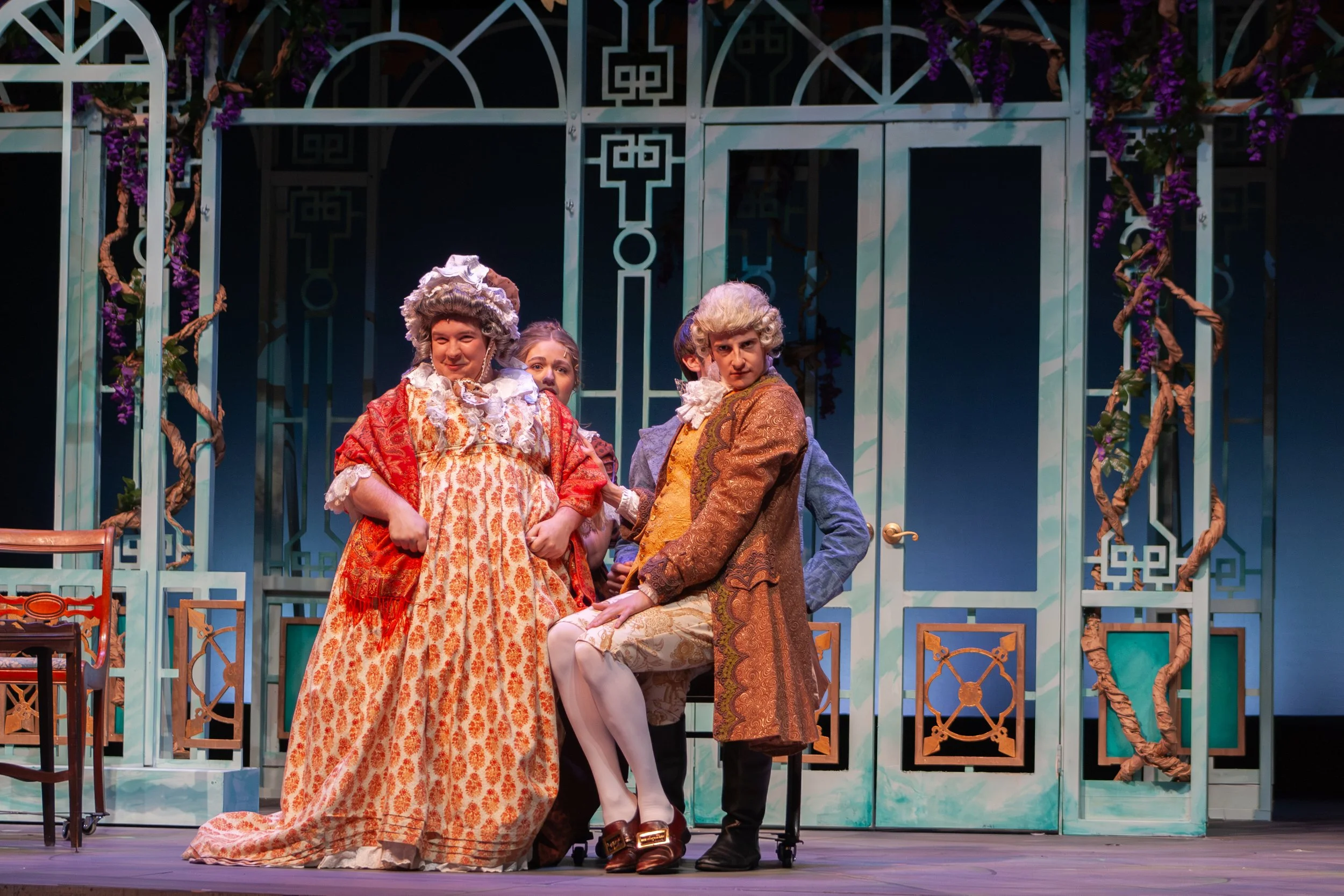 Four actors dressed in 18th-century costumes on stage, with a decorative garden-like backdrop featuring purple flowers and vines.