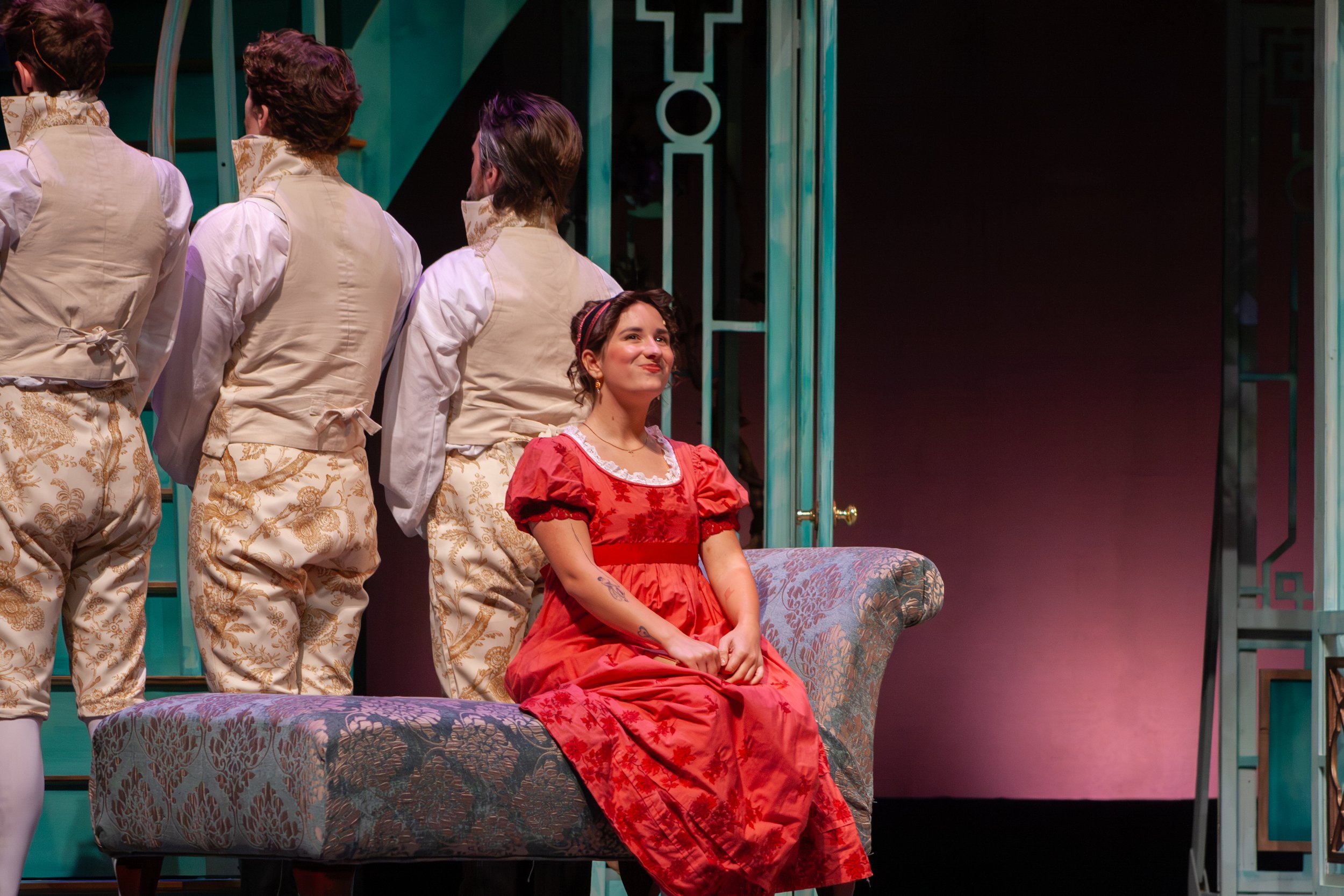 A woman with dark hair, dressed in a red gown, is sitting on a floral upholstered bench, smiling softly. Behind her, several men in similar beige and gold costumes are standing, with their backs to the camera. The setting appears to be a stage with a