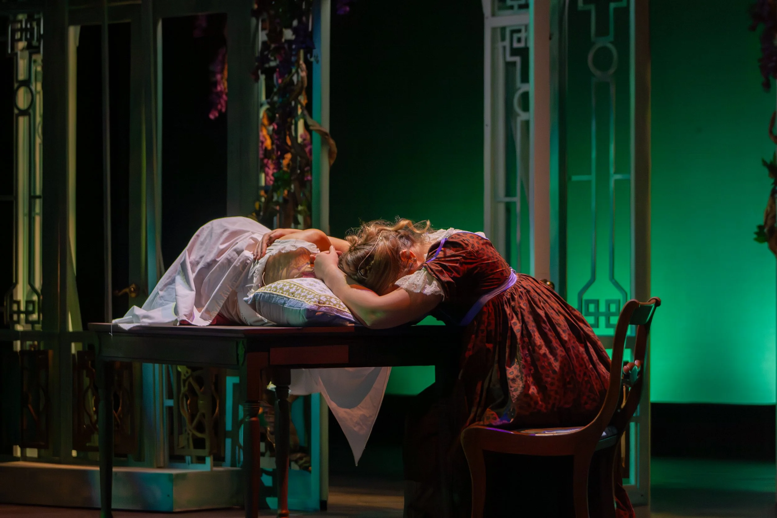 Two women wearing period costumes leaning on a table, with one woman resting her head on the other's shoulder, in a room with decorative green walls and furniture.