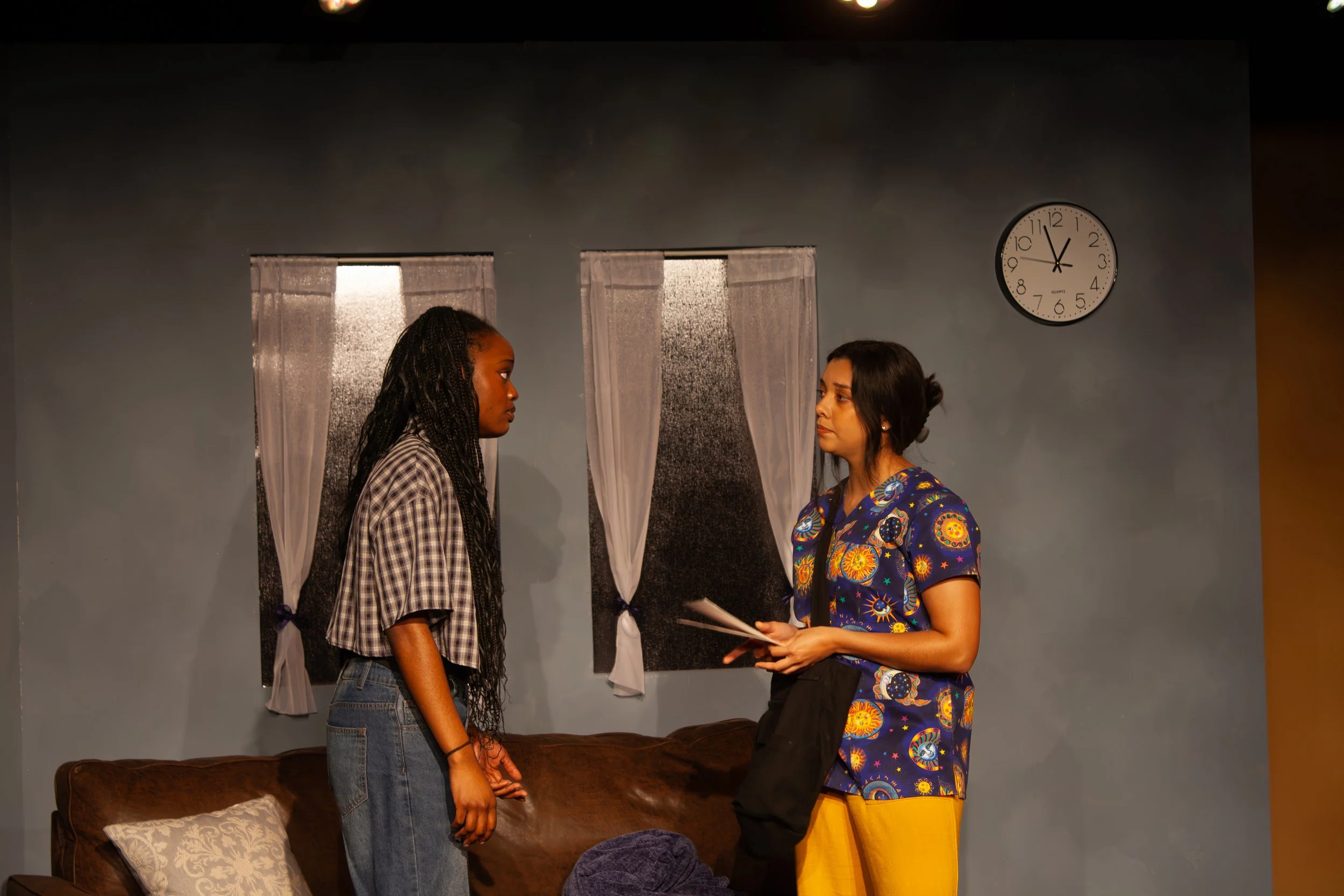 Two women facing each other indoors, with a dark gray wall and two windows covered with sheer white curtains behind them. A wall clock shows 11:55. One woman is wearing a checkered shirt and jeans, and the other is dressed in a colorful, patterned to