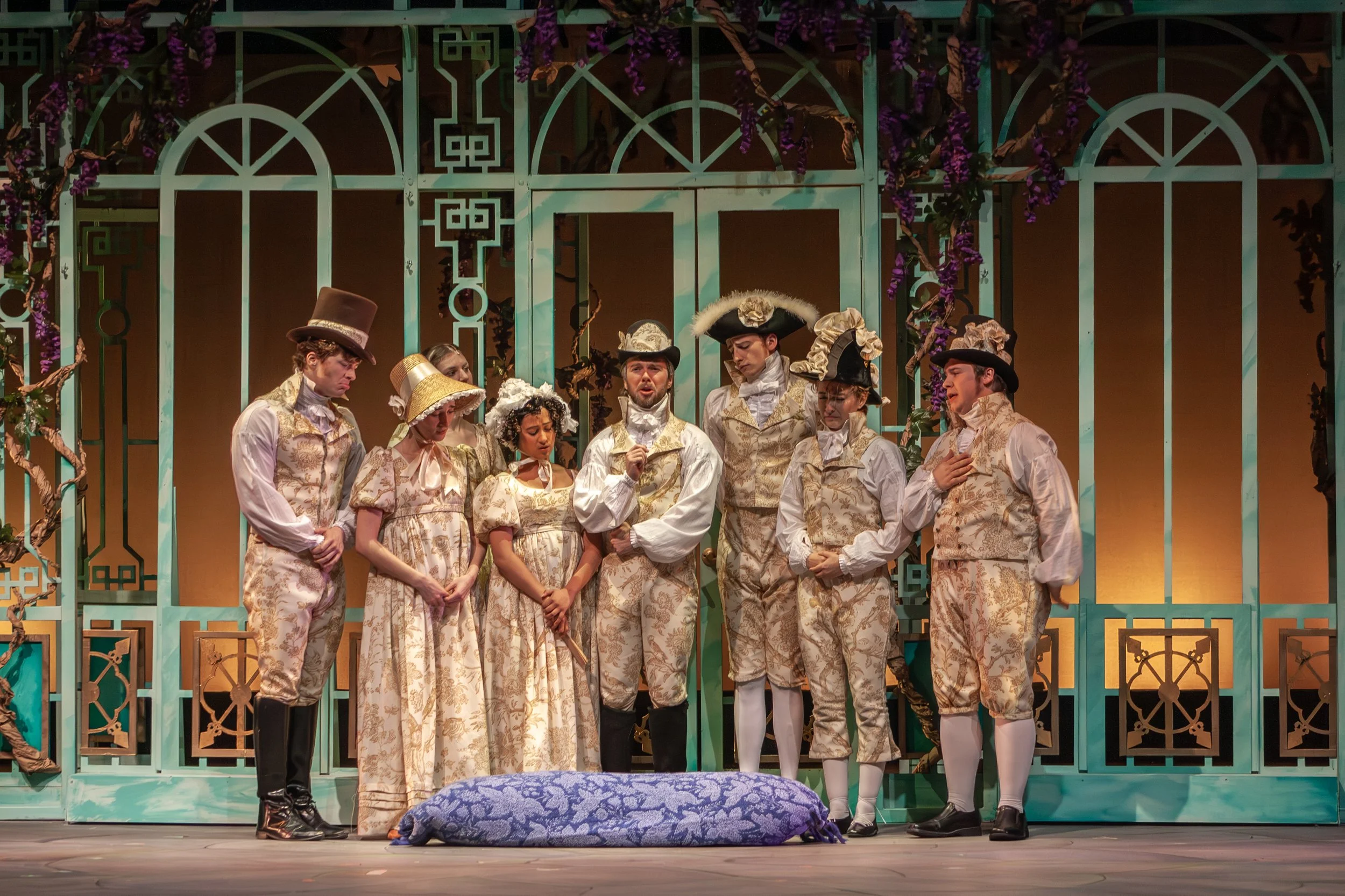 A theatrical stage scene with nine actors in period costumes, standing in front of a decorated set with teal framework and purple flowers. The actors are wearing ornate, floral-patterned clothing and hats, engaged in a dramatic moment.