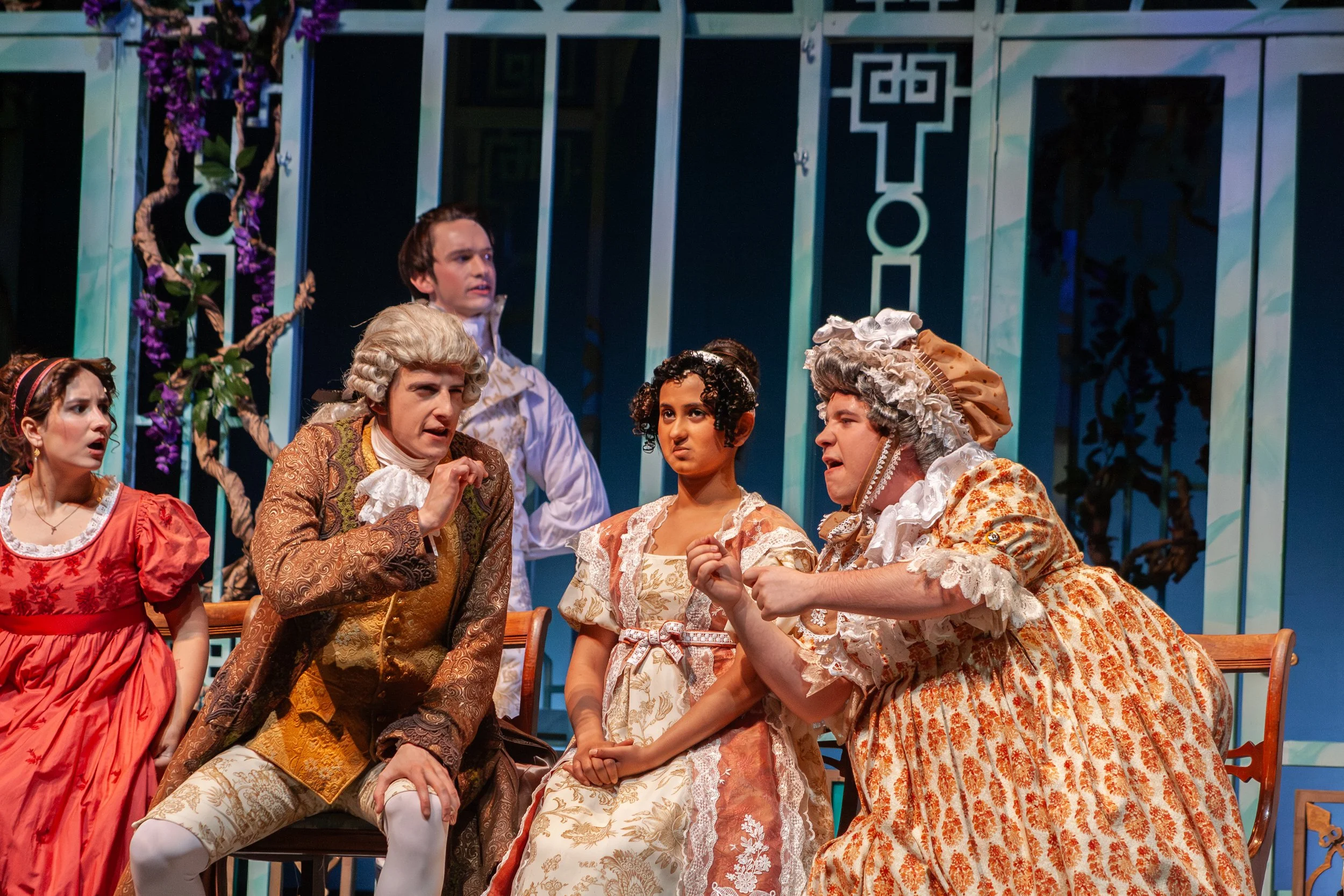A theatrical scene with six actors dressed in 18th-century costumes, including elaborate dresses and wigs, performing on stage with a decorative backdrop featuring a tree with purple flowers and geometric patterns.