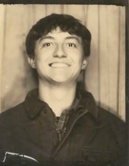 Black and white photo of a young man smiling with a slight twist of his head, standing in front of a curtain.