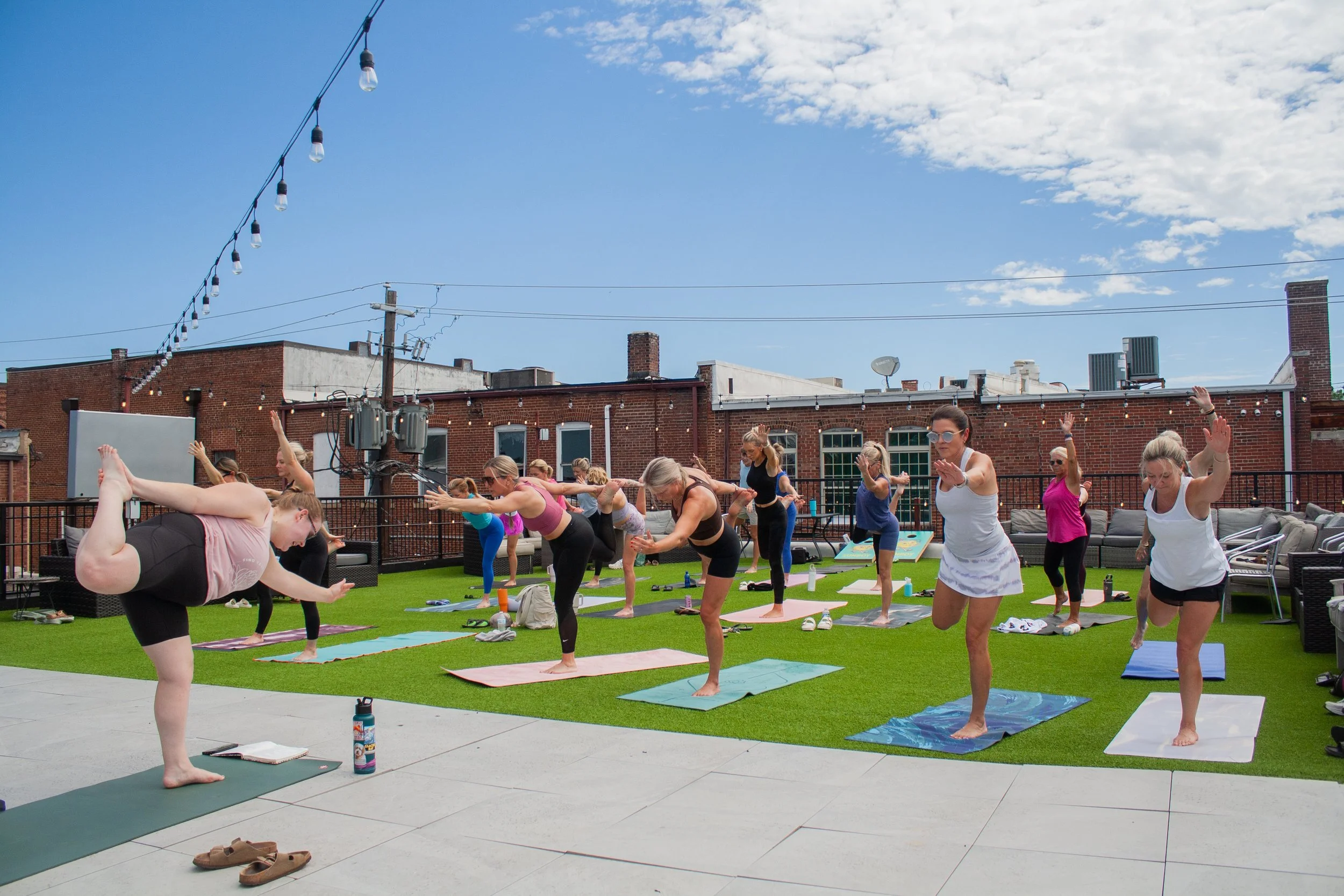 People participating in an outdoor yoga class on a rooftop terrace, practicing yoga poses on mats with city buildings and string lights in the background on a partly cloudy day.