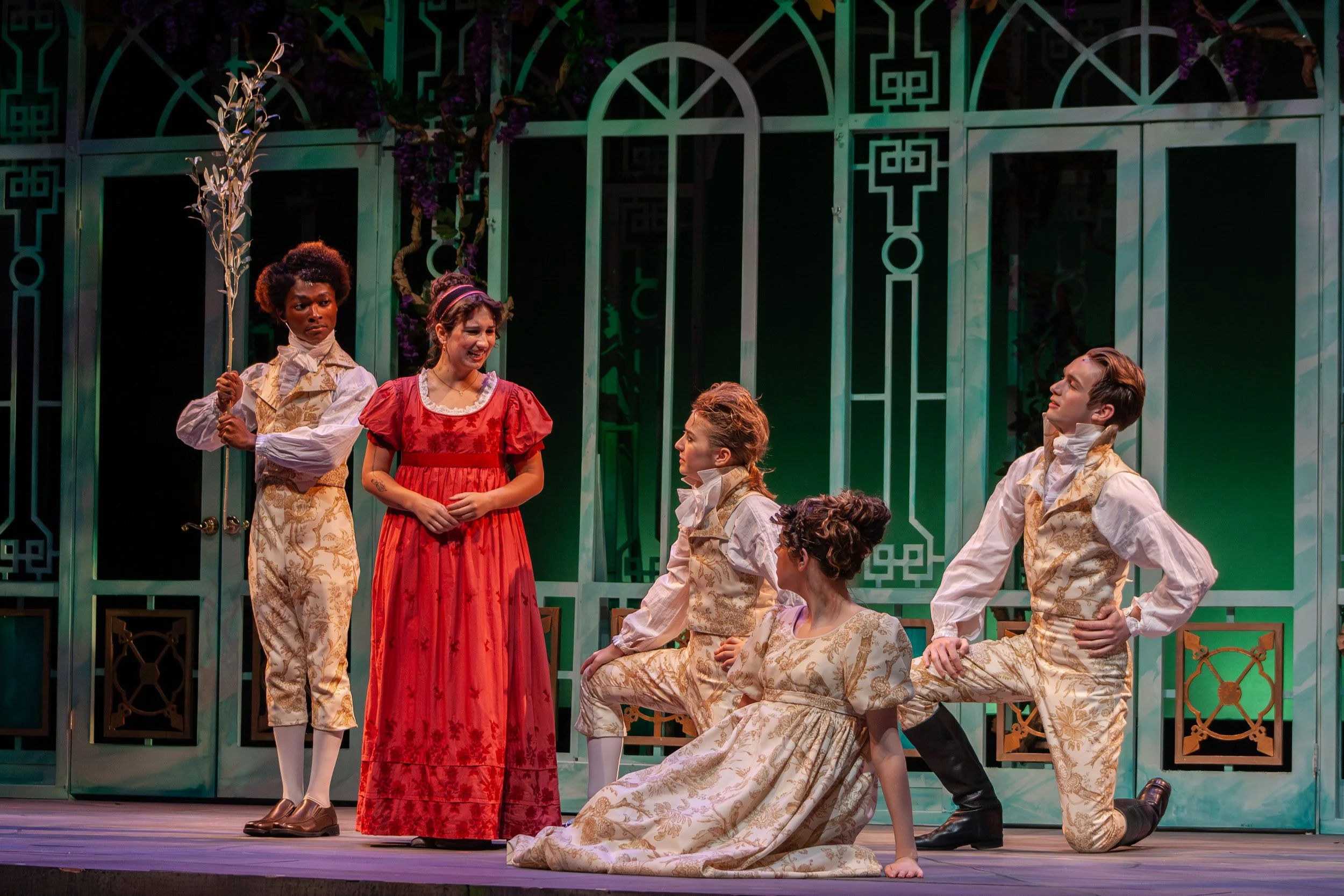 A scene from a theatrical performance with six actors in period costumes on stage. The backdrop is a teal-colored set with geometric patterns and plant decorations. One woman in a red dress is smiling and standing in the center, while others are enga