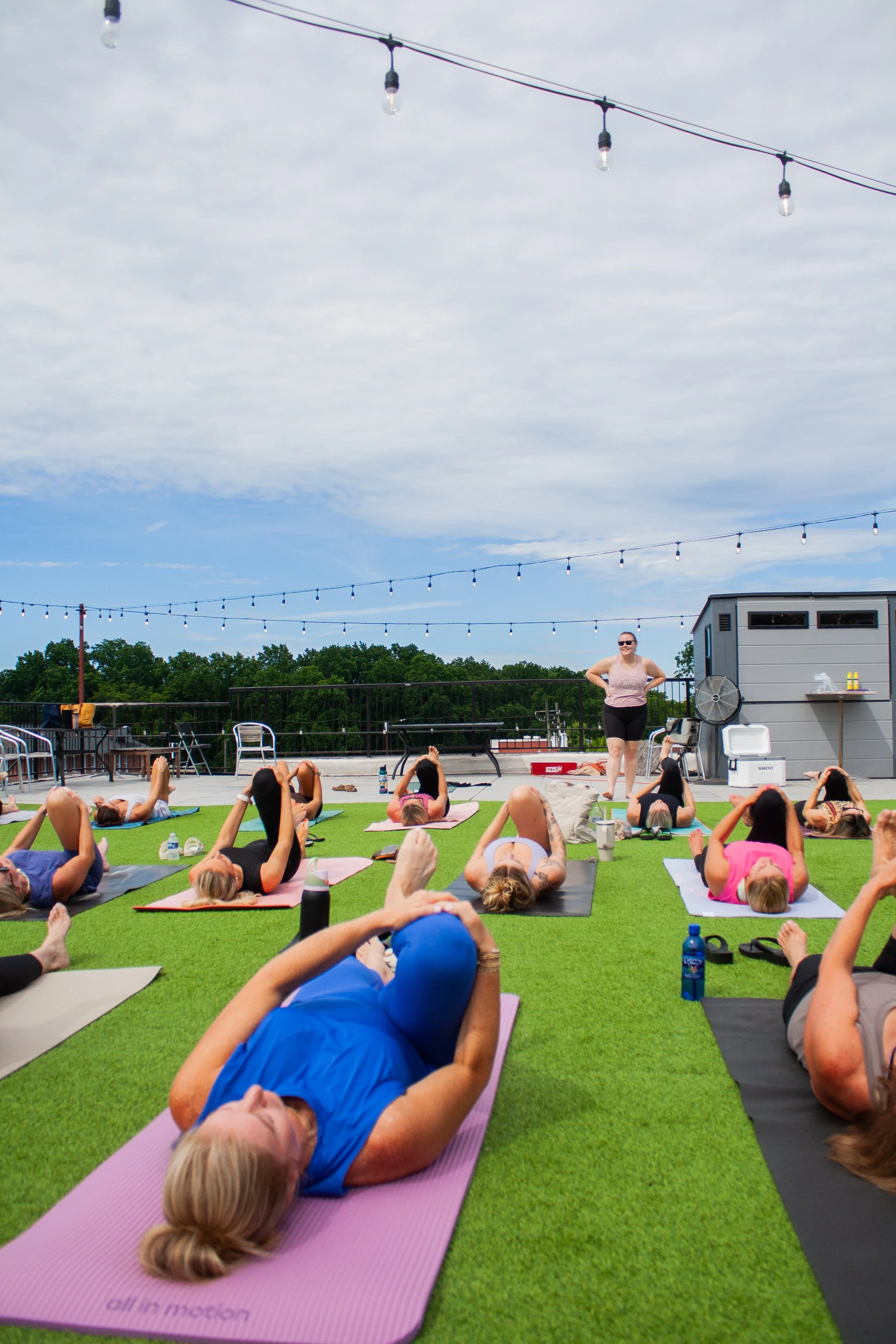 A group of people participating in an outdoor yoga class on a rooftop, lying on colorful yoga mats on artificial grass, with string lights overhead and a partly cloudy sky. A female instructor is standing and guiding the class.