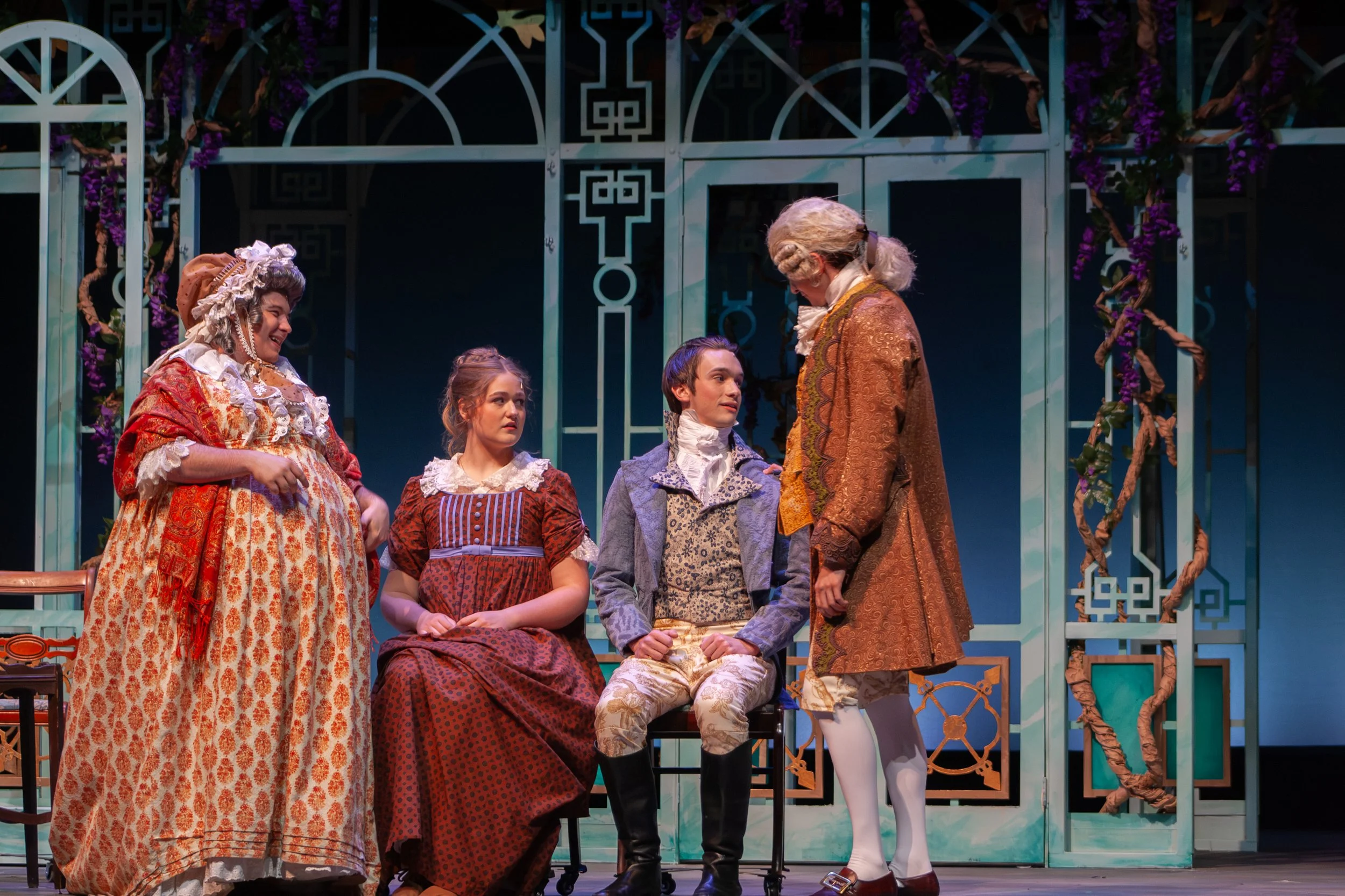 Actors dressed in 18th-century period costumes on stage, engaging in a theatrical scene with a decorative background featuring blue metal arches and purple flowers.