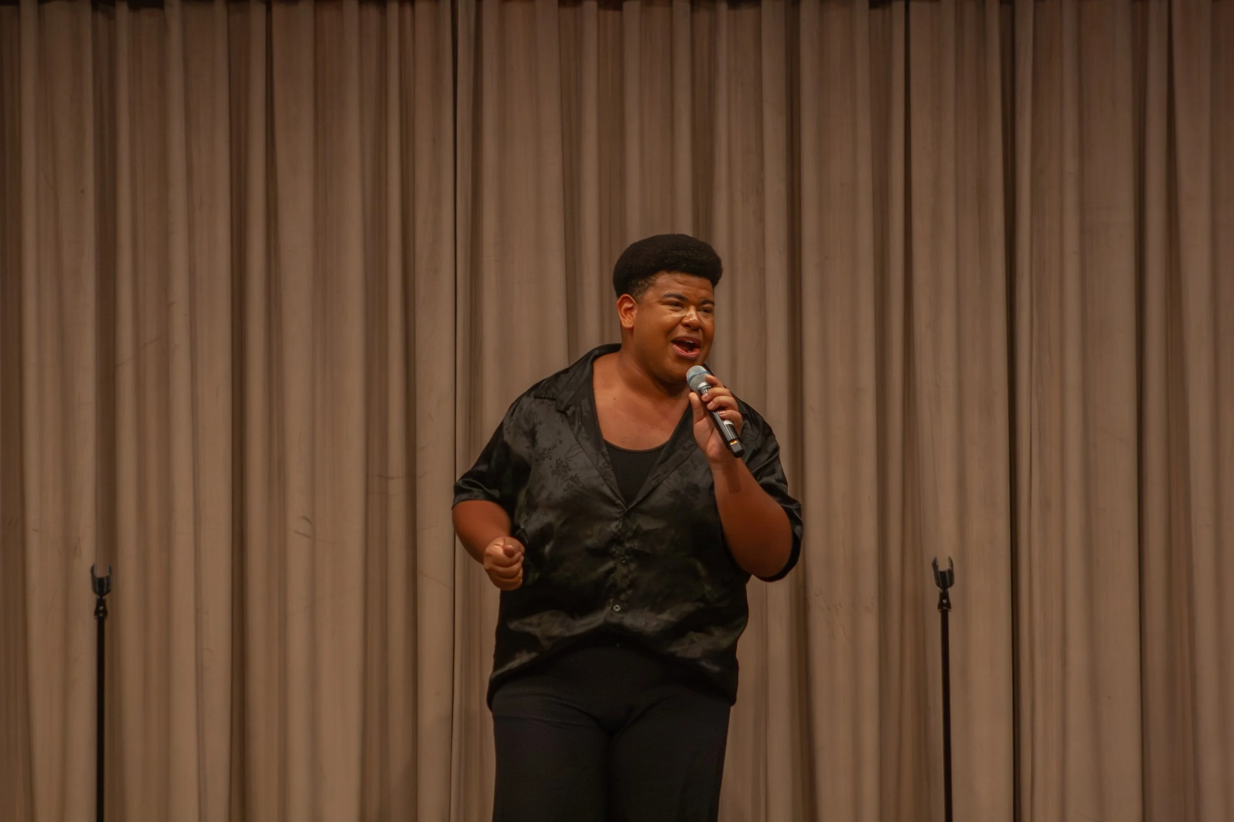 A woman singing into a microphone on stage with a beige curtain backdrop.