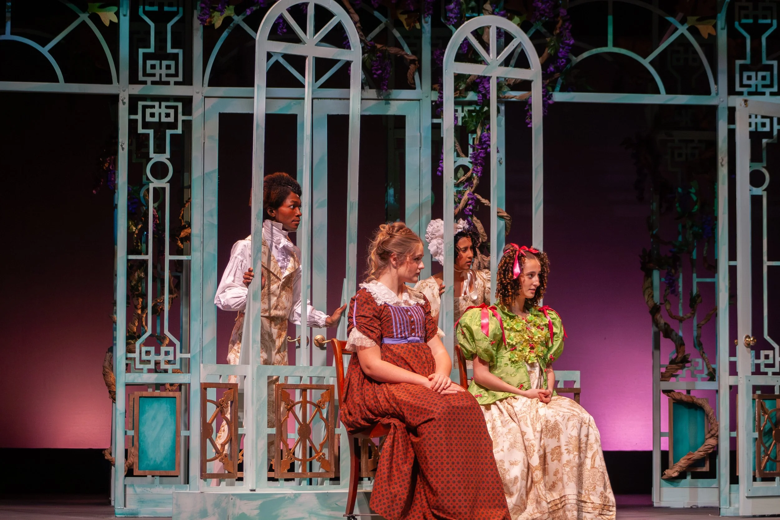 Scene from a theatrical play featuring five women dressed in vintage costumes, sitting and standing behind a decorative white and pastel-colored metal fence with purple flowers.