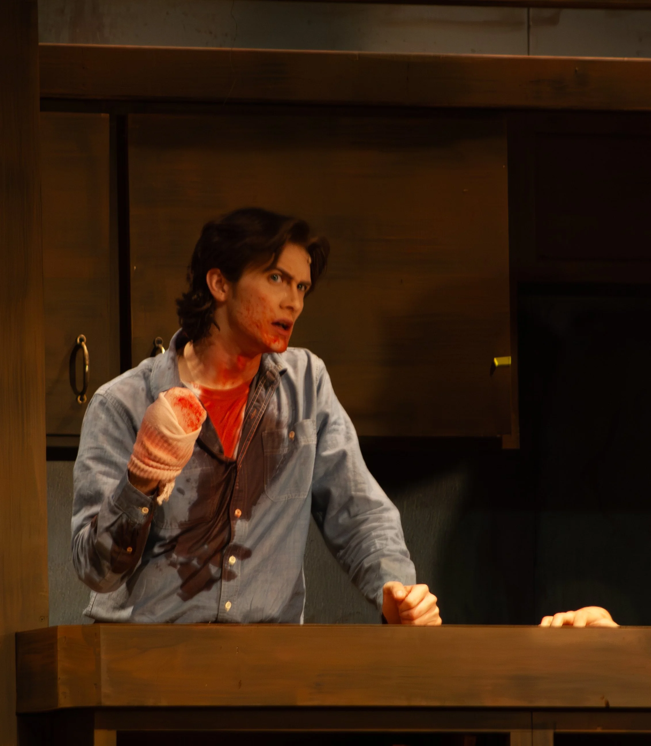 A young man with brunette hair and a serious facial expression is wearing a light denim shirt and a brown leather apron, sitting at a wooden counter in a dimly lit room. His face, neck, and hand are covered in red paint or a similar substance. The ba