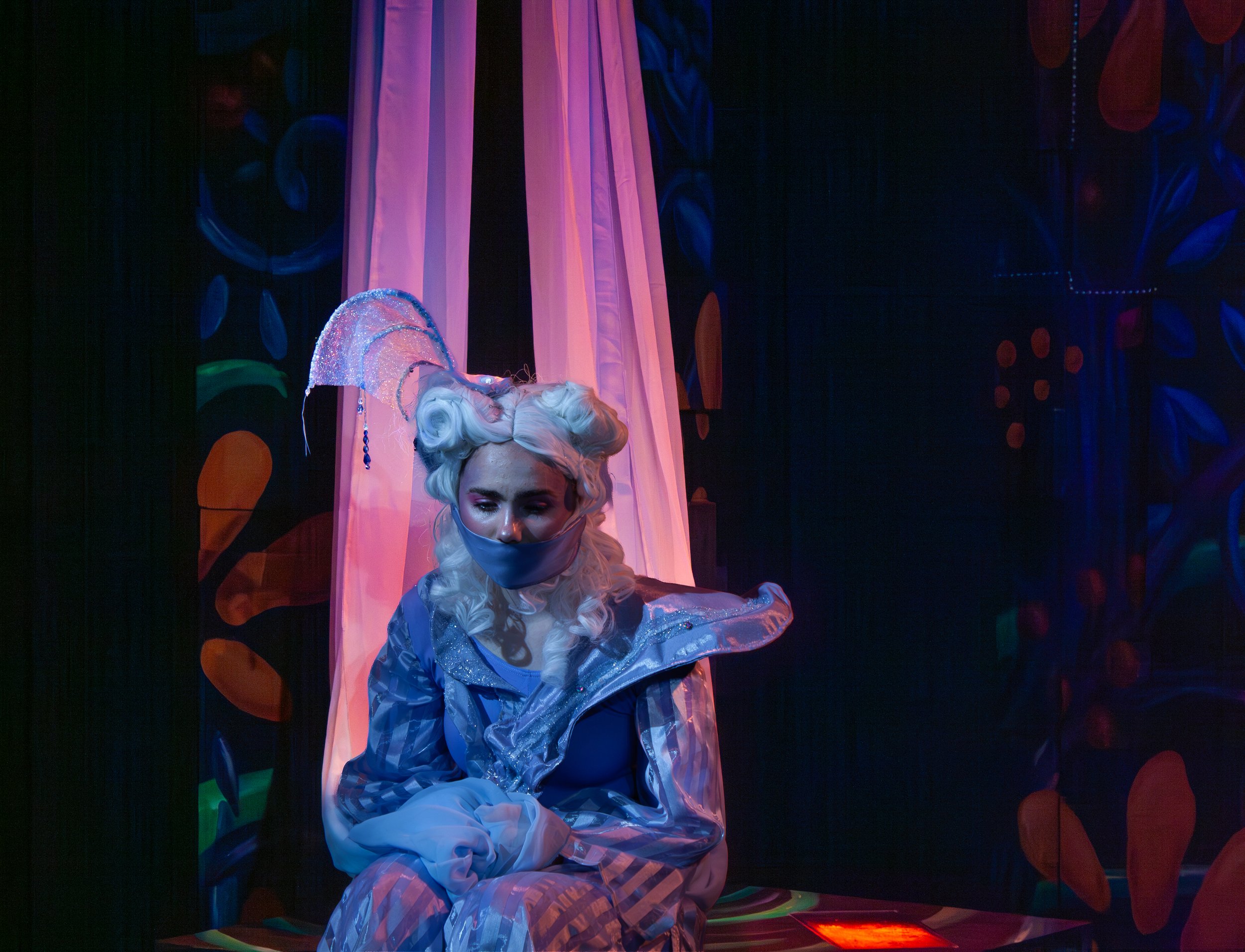 A woman with platinum blonde hair wearing a sparkly, elaborate outfit, sitting on a stage with dark background and colorful decorations, wearing a face mask.