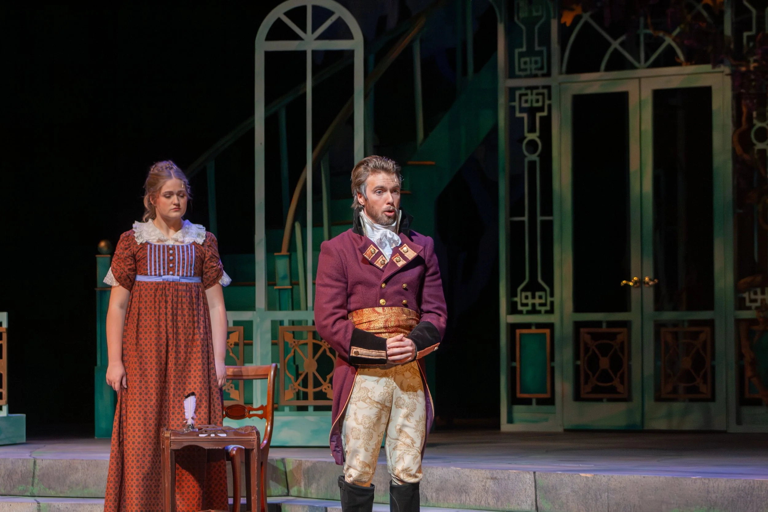 Three actors on stage in period costumes performing in a play. The man in the center is dressed in an 18th-century style coat and ornate breeches, standing with his hands clasped. The woman on the left is in a long patterned dress with lace details, 