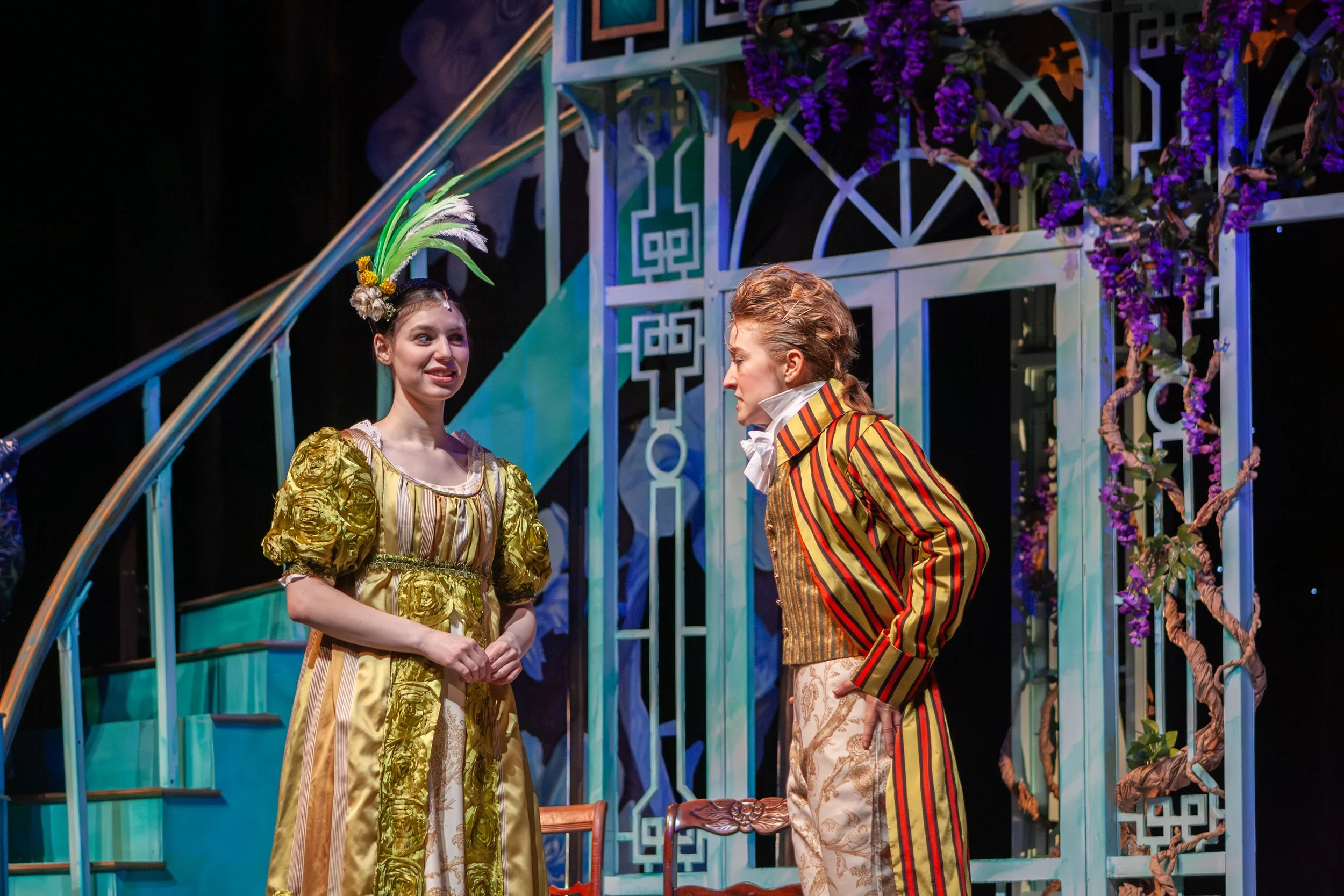 Two women in period costumes engaged in a theatrical scene on stage with a decorative set, including purple flowers and intricate architectural elements.