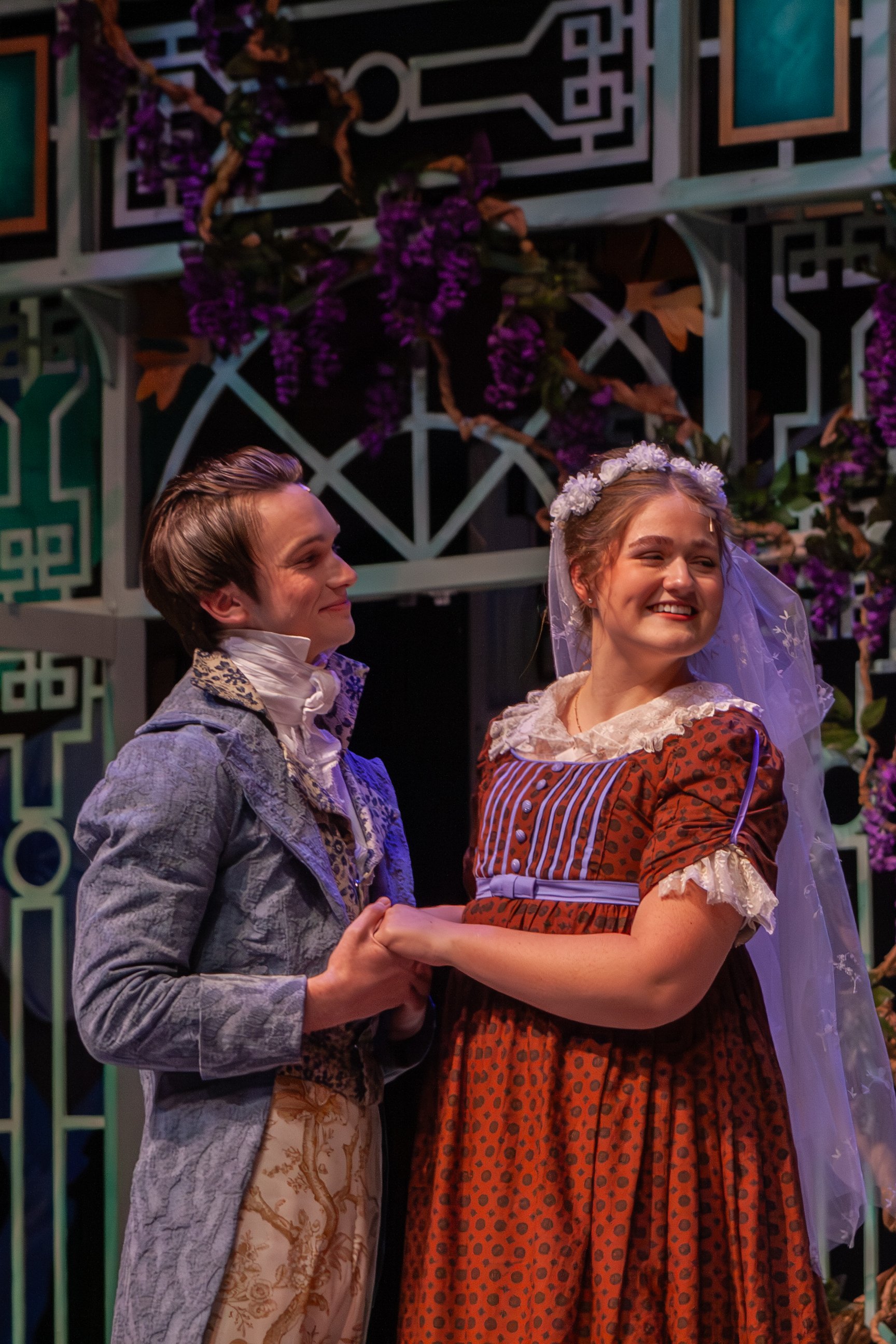 Two young people dressed in period costumes are holding hands and smiling, with a floral and decorative background.