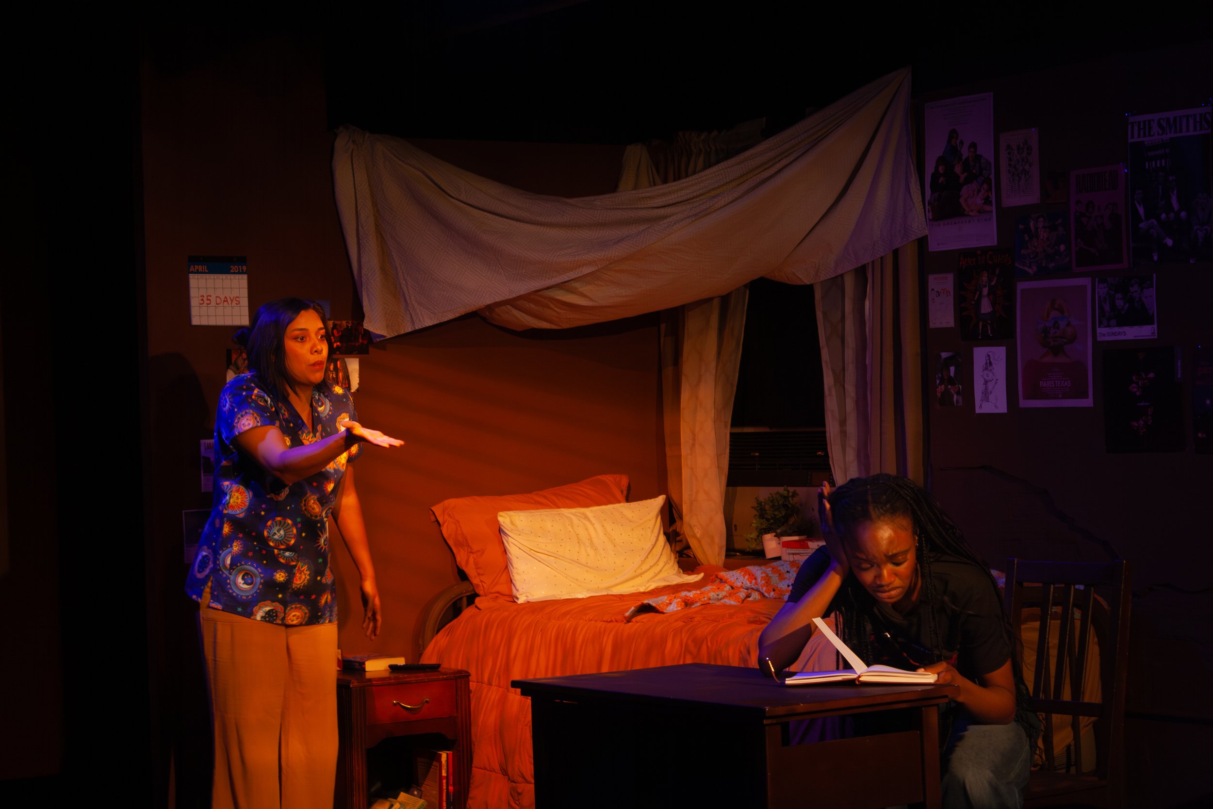 Scene from a theatrical play or performance set in a bedroom, featuring a woman in a colorful shirt gesturing, and another woman sitting and writing in a notebook, with decorated walls and furniture.