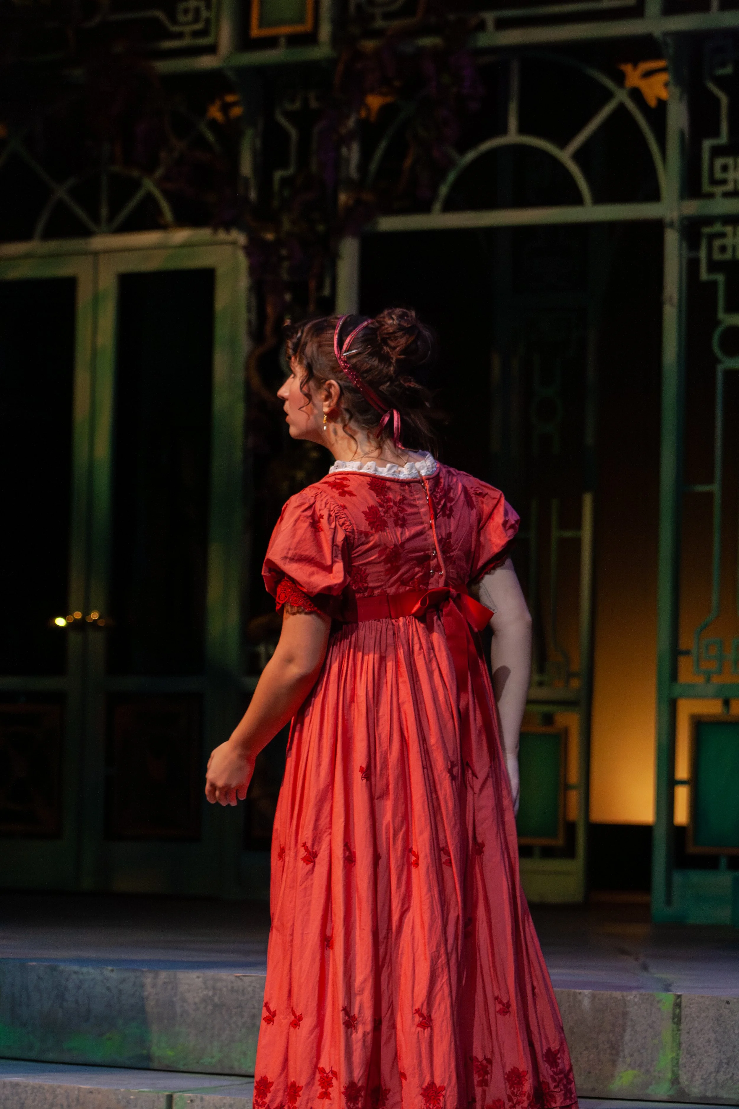 A woman wearing a long red dress with floral embroidery and a white lace collar stands on stage in front of a decorative stage set, with her hair styled in curls and adorned with ribbons.