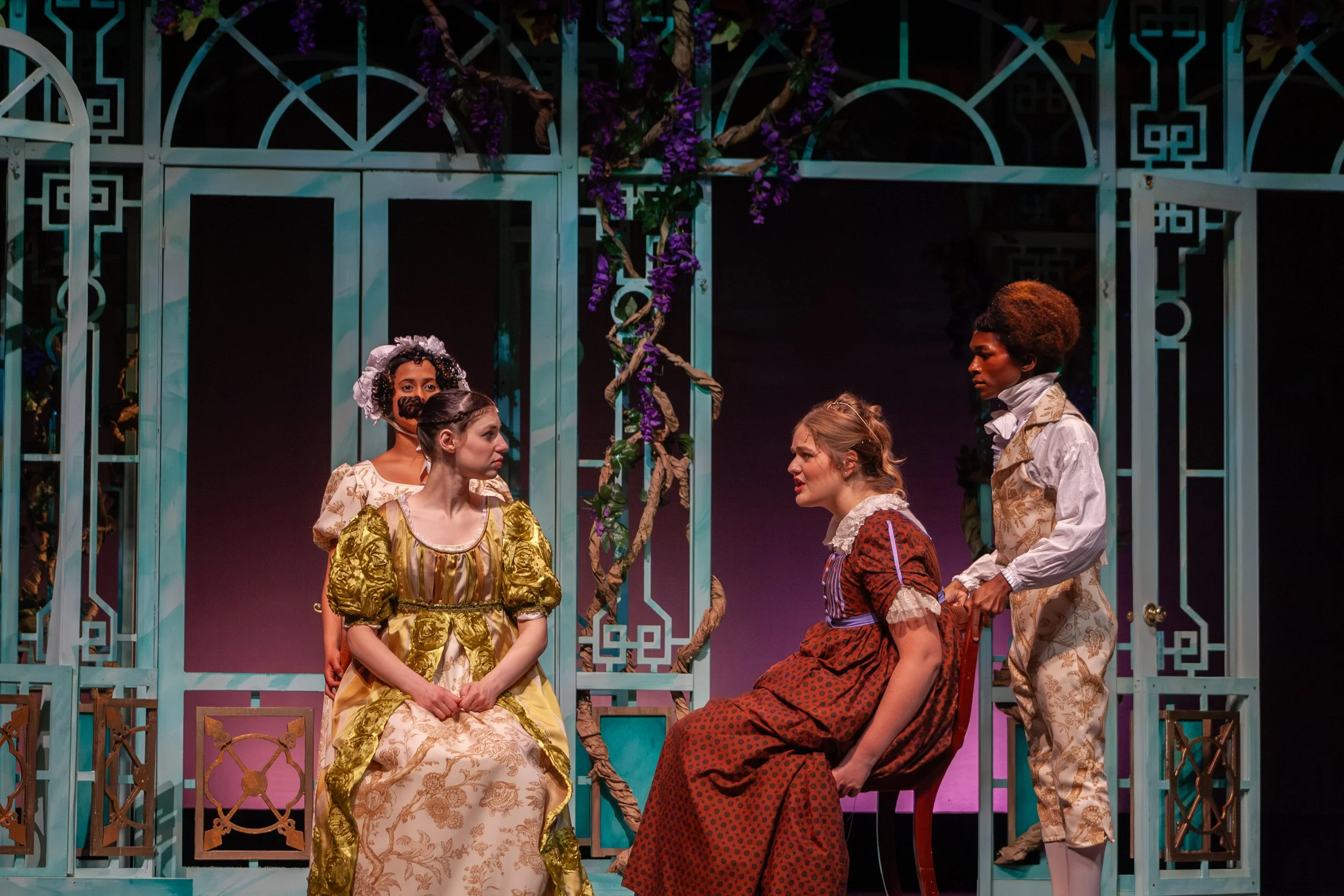 The image shows a theatrical scene with five actors dressed in period costumes. They are positioned in front of a decorative, teal-colored lattice structure with pink and purple flowers and twisted branches. The actors appear to be engaged in a drama