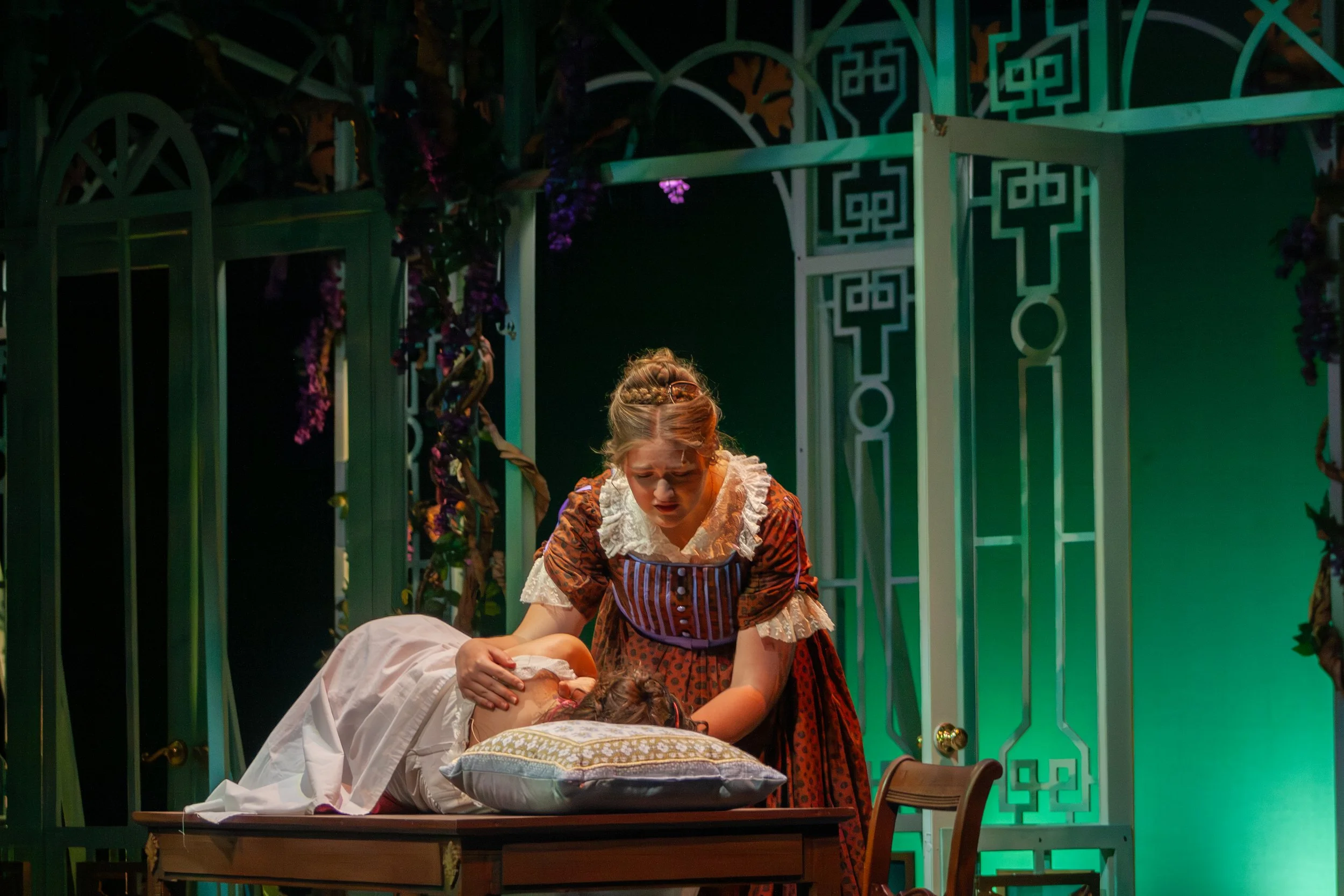 A theatrical scene with a woman in a historical costume leaning over a person lying on a table, with a green-lit set and decorative panels in the background.
