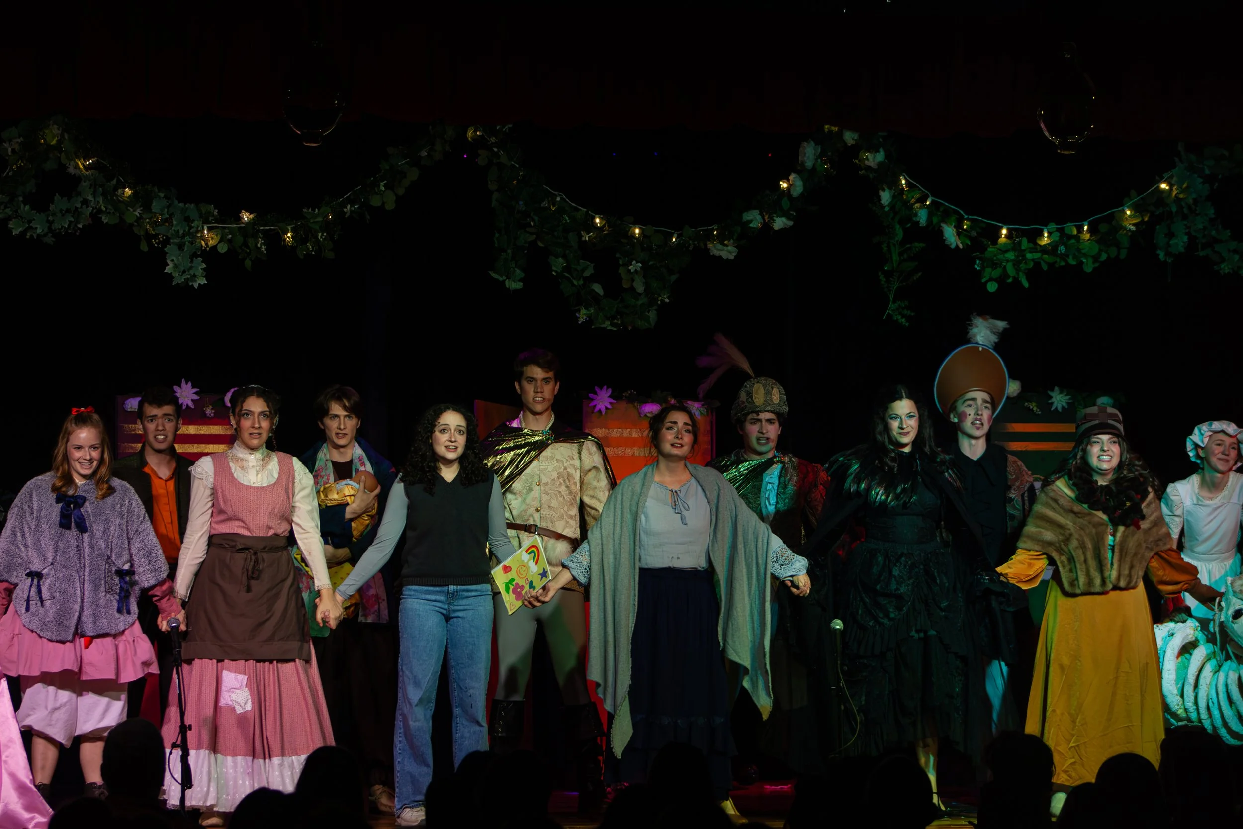 A group of people dressed in theatrical costumes standing on stage after a performance with decorative lights and greenery hanging above them.