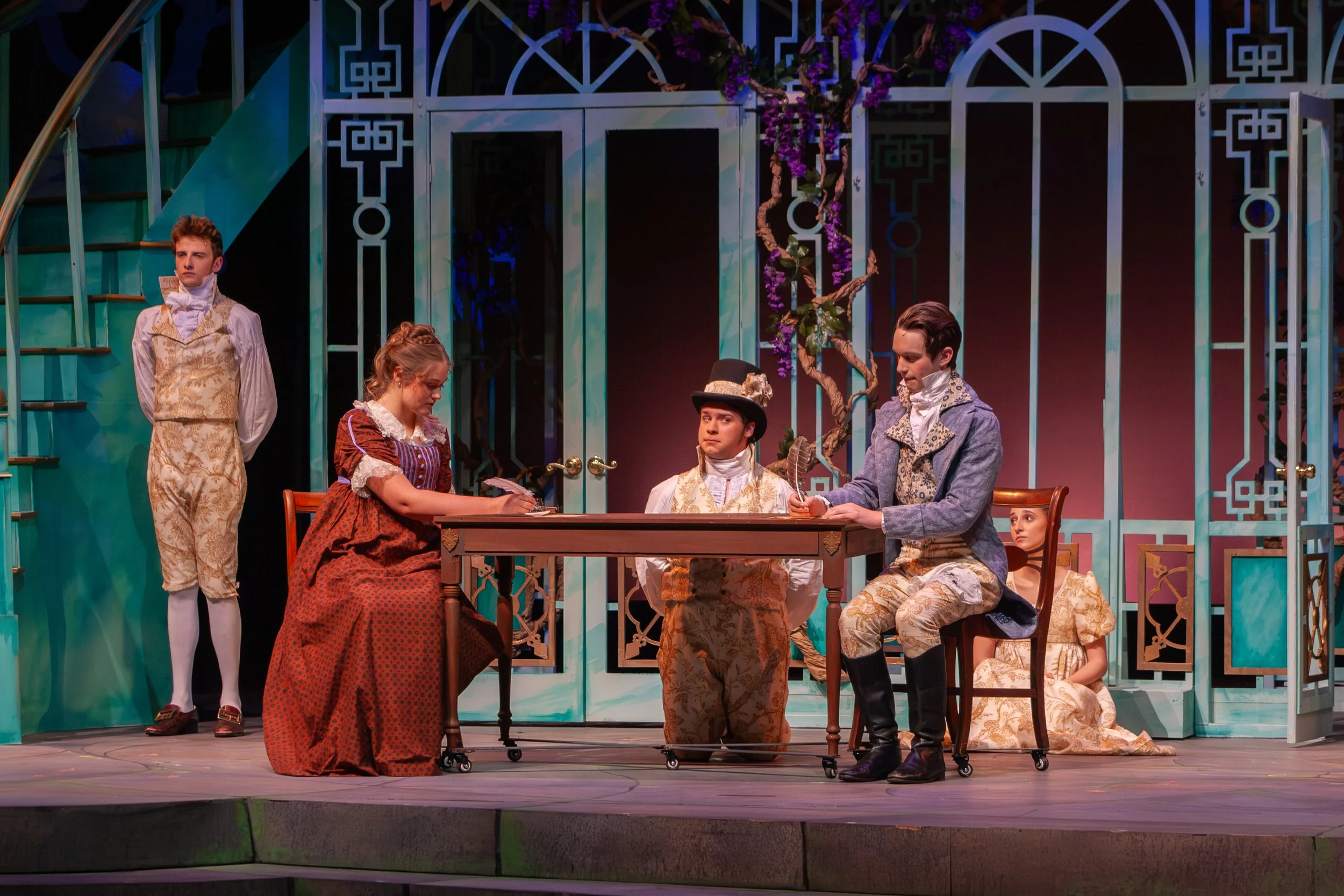 The scene shows actors in period costumes performing on stage, with some standing and others sitting around a table, inside a set designed like a decorative garden with teal gates and purple flowers.
