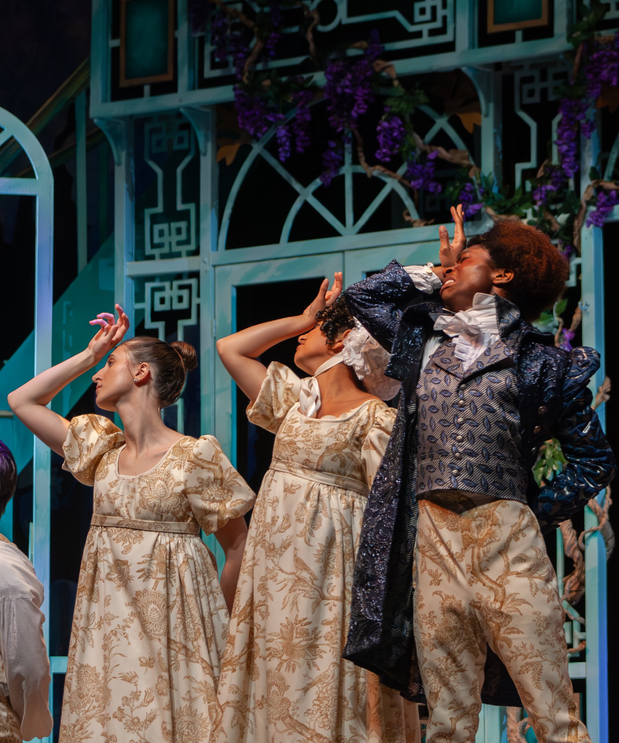 A theatrical scene with four women in vintage-style dresses, one woman with a dark curly coat, all engaged in expressive gestures against a background of a decorative balcony and purple flowers.