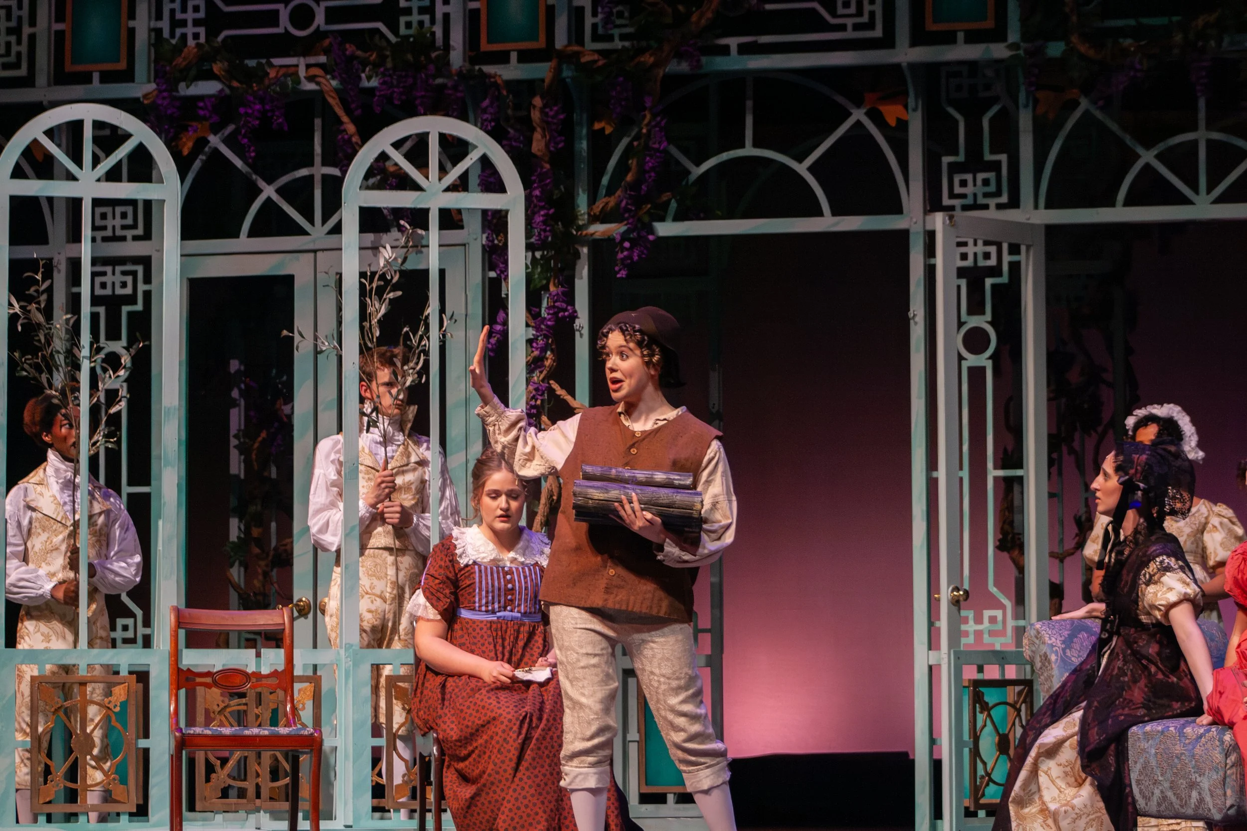 Actors on stage performing in costumes from the play 'The Enchanted Pig', surrounded by a decorative set with garden elements.