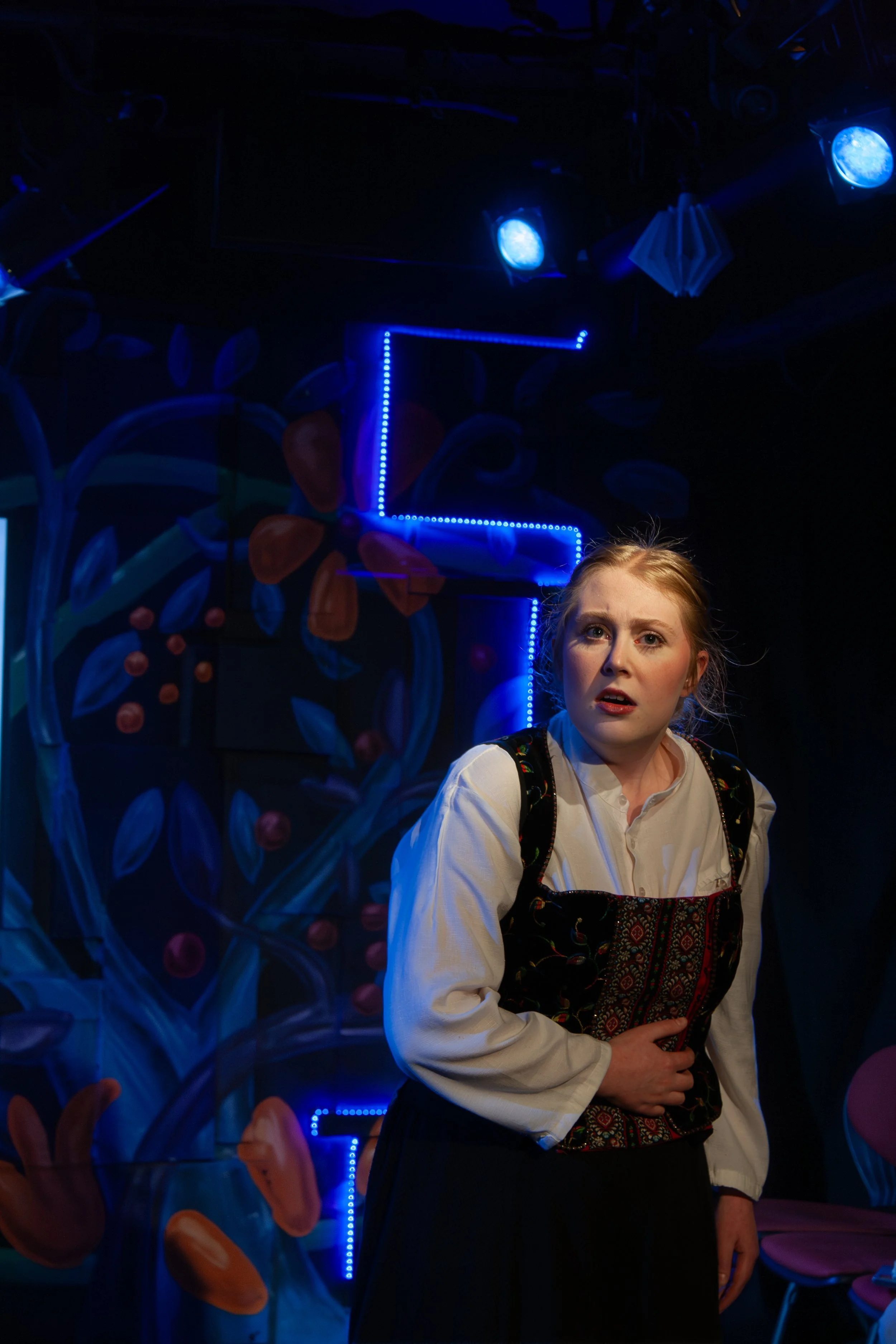 Young girl with red hair, wearing traditional dress, standing indoors in front of a colorful mural with floral designs and neon blue lights, looking slightly surprised.