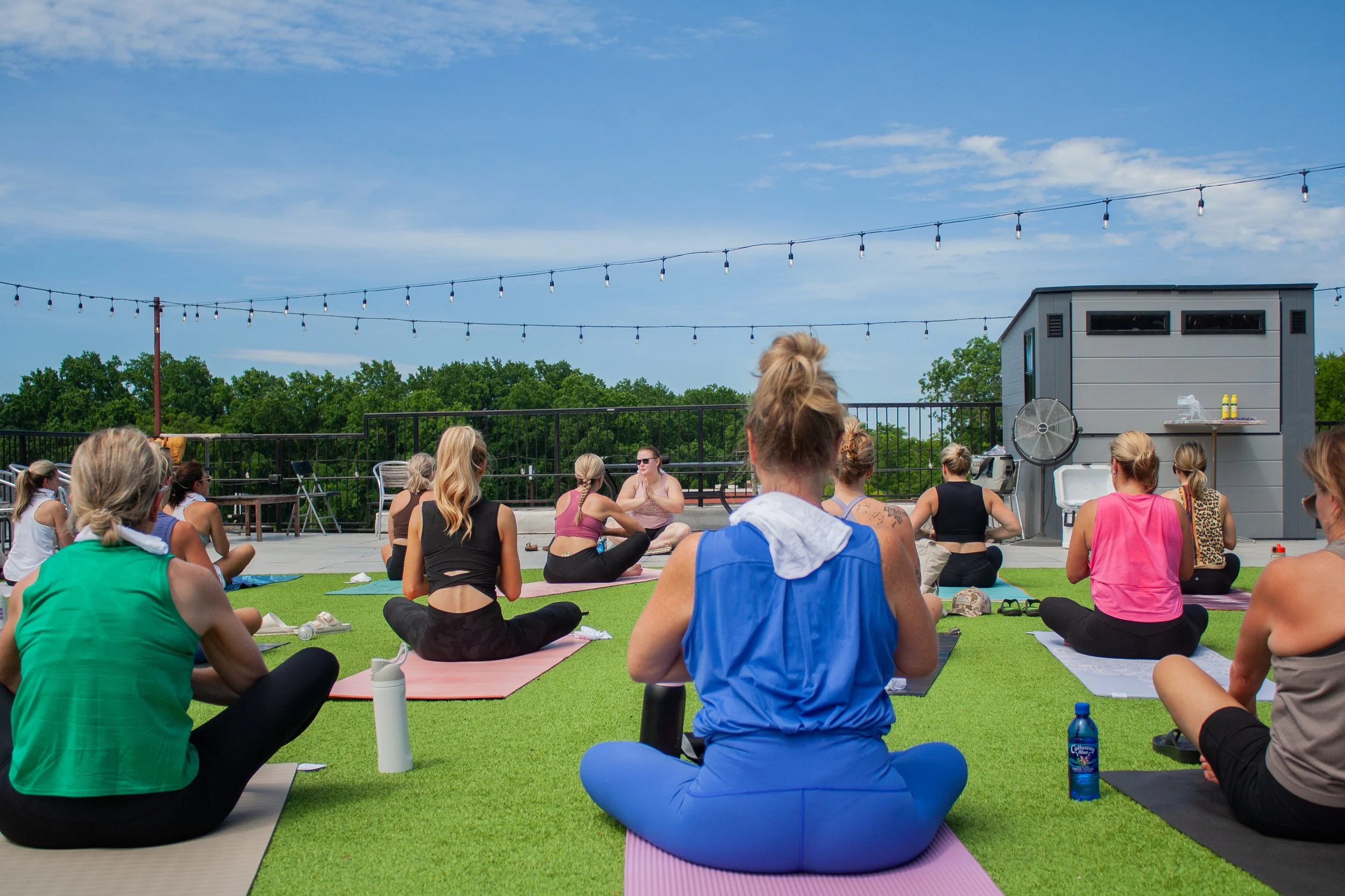 Group of people practicing yoga outdoors on a rooftop under string lights, with a small building and trees in the background.