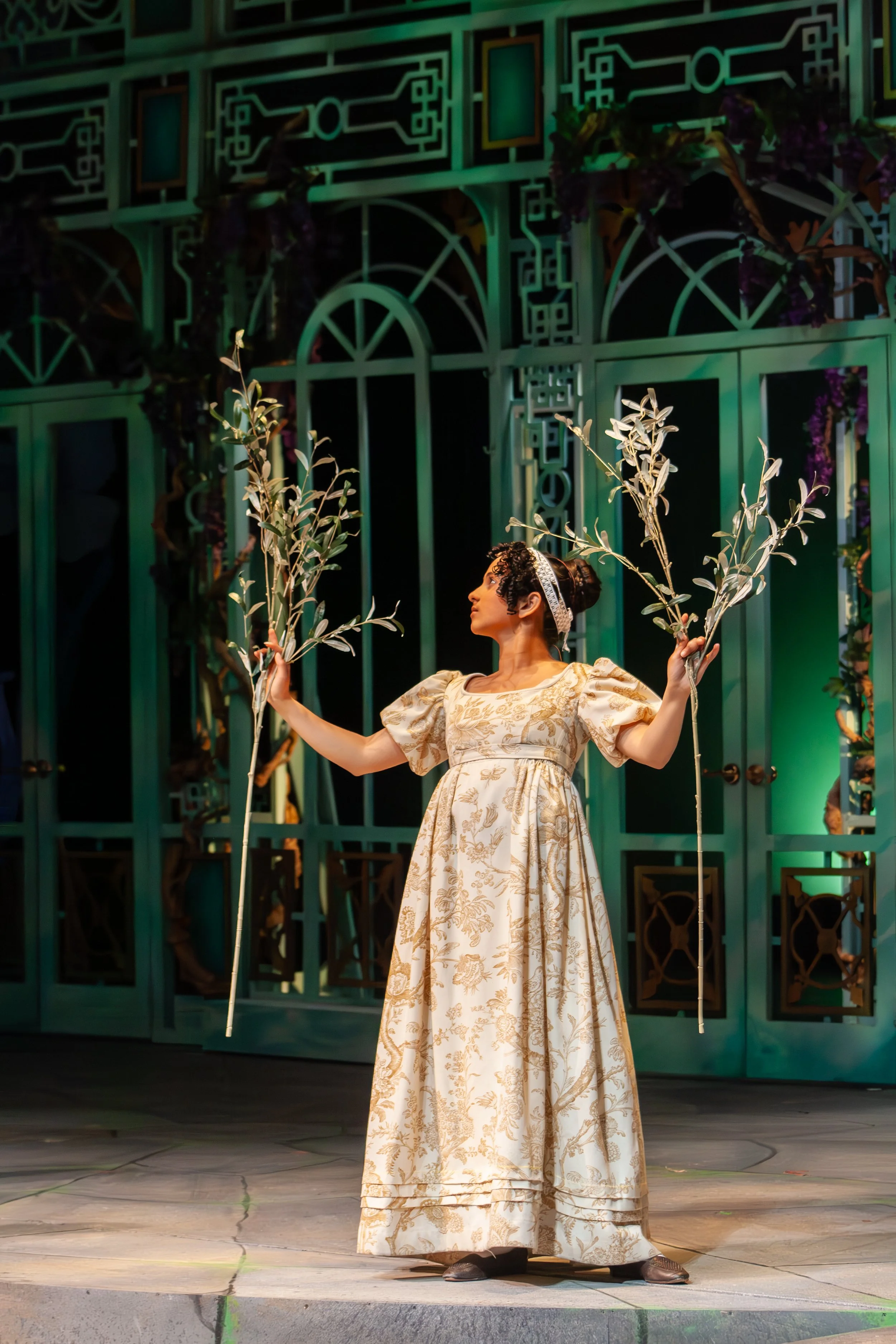 A woman in a long, ornate, gold-patterned dress stands on stage, holding two scepters with leafy branches, looking upward. She is wearing a silver tiara and has her hair styled in an updo. The background features a large, decorative green structure w