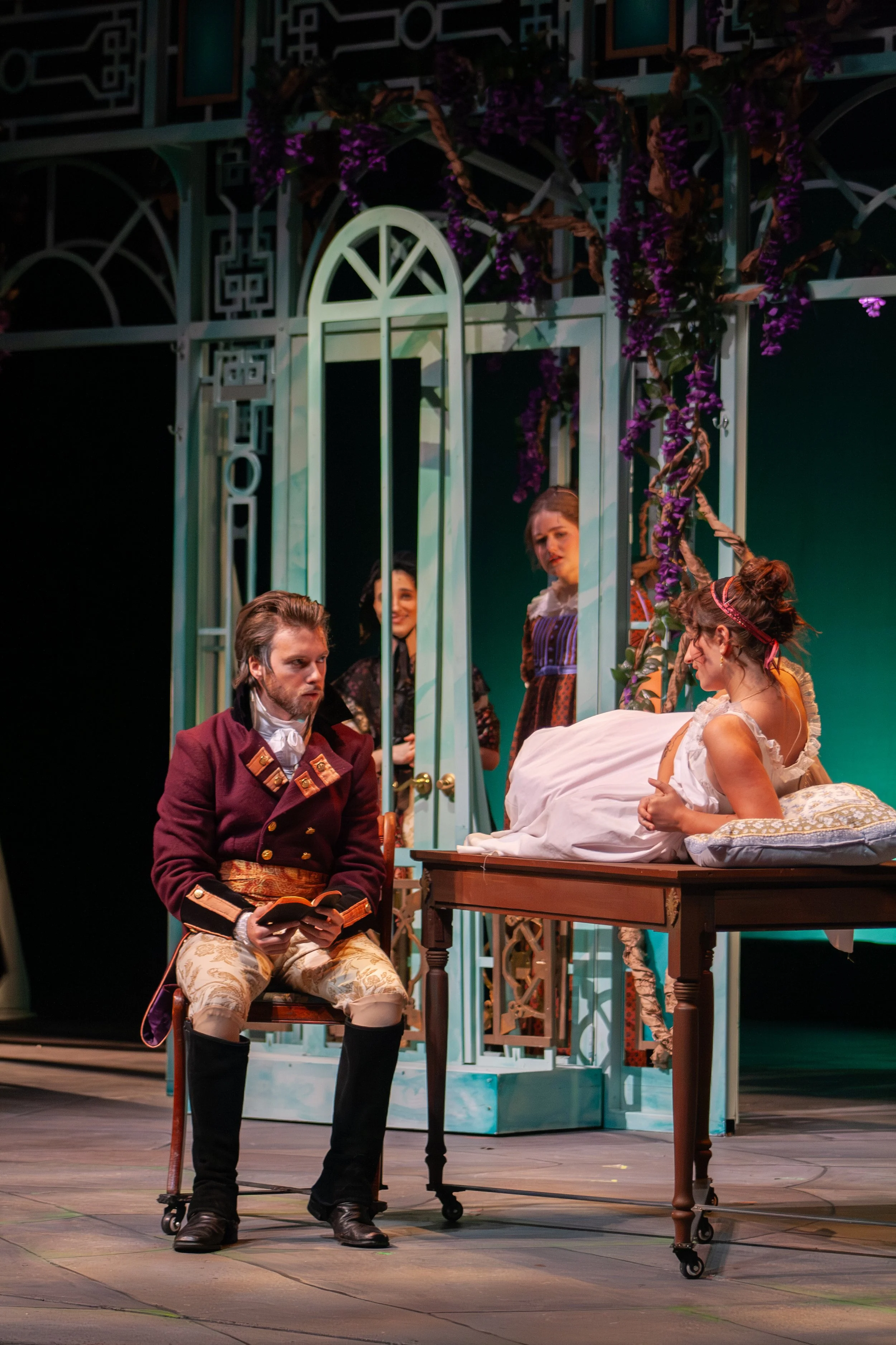 Scene from a theatrical play with actors in period costume, including a man in a maroon coat and beige pants, and women in dresses, with a decorative set featuring a large white window frame, purple vines, and flowers in the background.