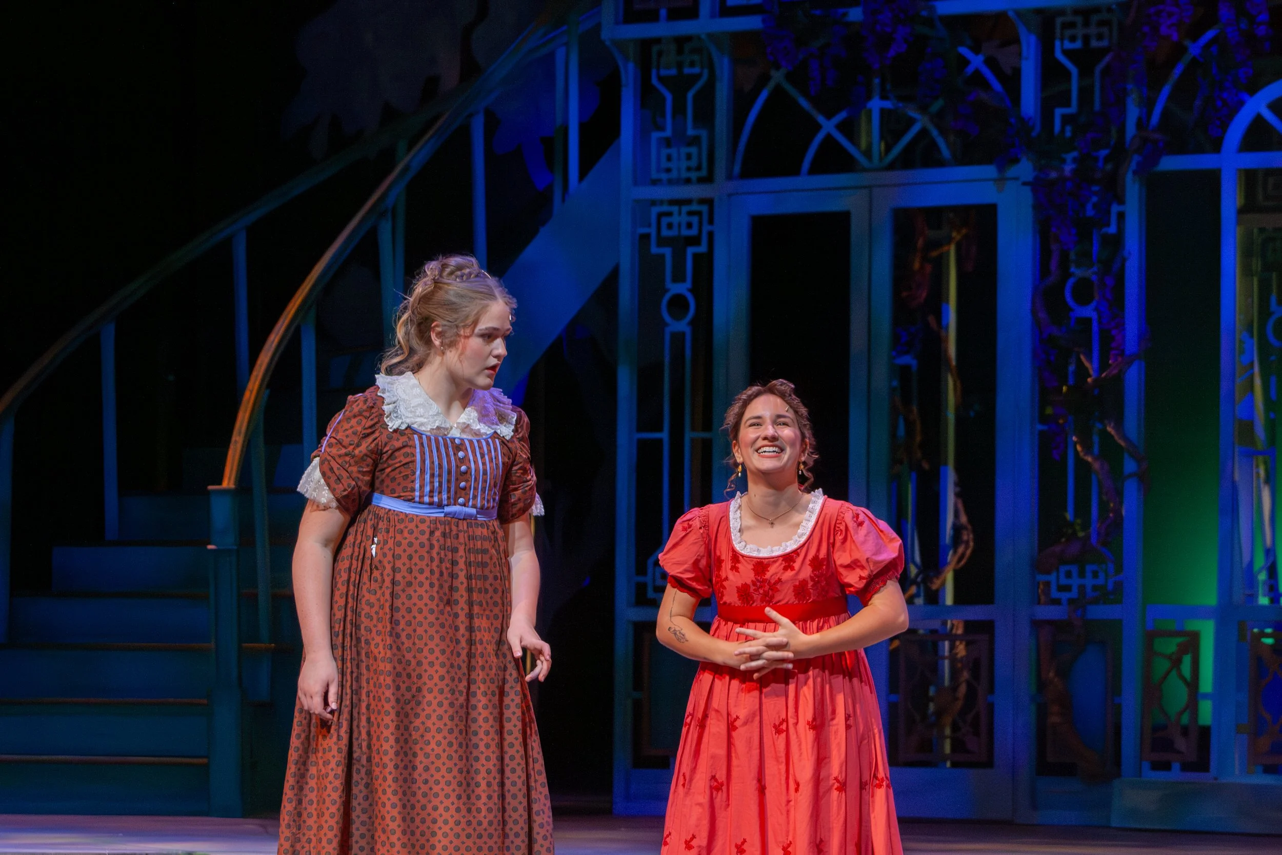 Two women in period costumes standing on a stage with a decorative blue metal backdrop. One woman is wearing a brown patterned dress with lace collar, and the other is in a red dress with floral embroidery, both smiling and engaged in conversation.